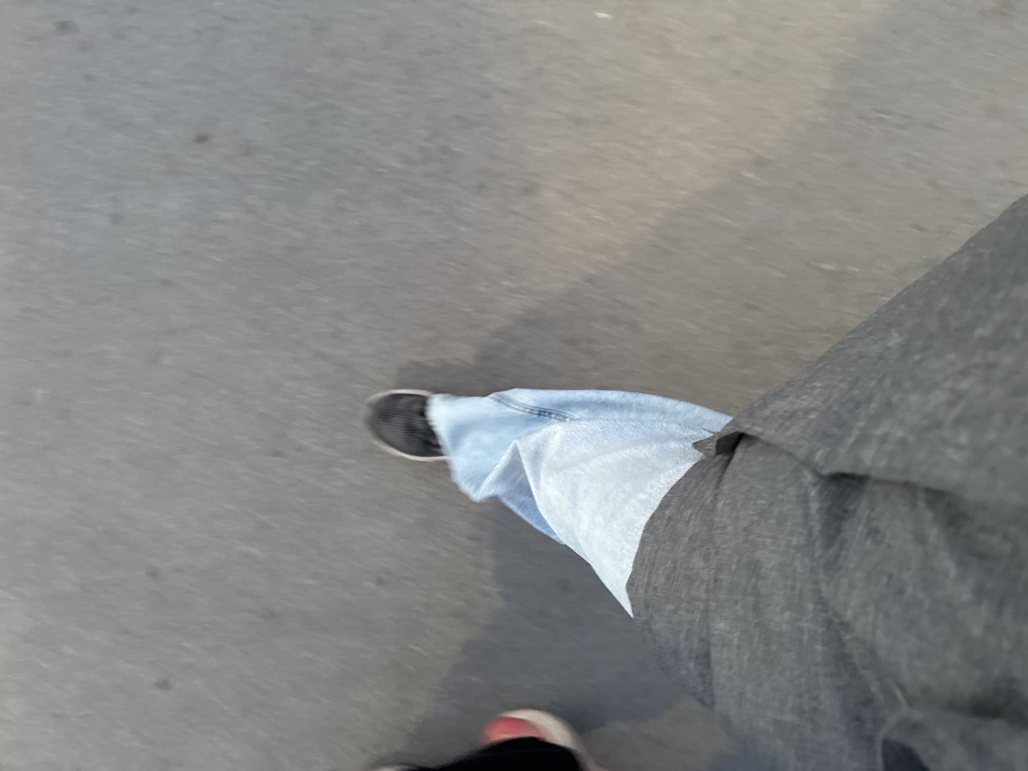 The image shows a first-person perspective of a person walking on a paved surface, likely a sidewalk or street. The person is wearing jeans with one leg partially covered by a dark gray garment, possibly a coat or jacket. Two shoes are visible: one is black and the other appears to have a red or pink accent. The background is a gray, textured surface, likely asphalt or concrete. The location in Berezniki, Russia cannot be confirmed from the image.