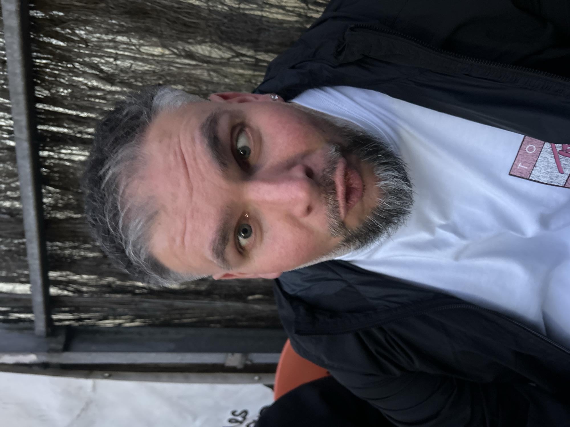 A close-up portrait shows a middle-aged man with graying hair and a salt-and-pepper beard looking directly at the camera. He has light skin and green eyes. He is wearing a white T-shirt with a graphic design visible on the chest and a black jacket. The background consists of a dark, textured wall or fence. The photo was taken outdoors. The location, Hem, France, cannot be confirmed with this image.