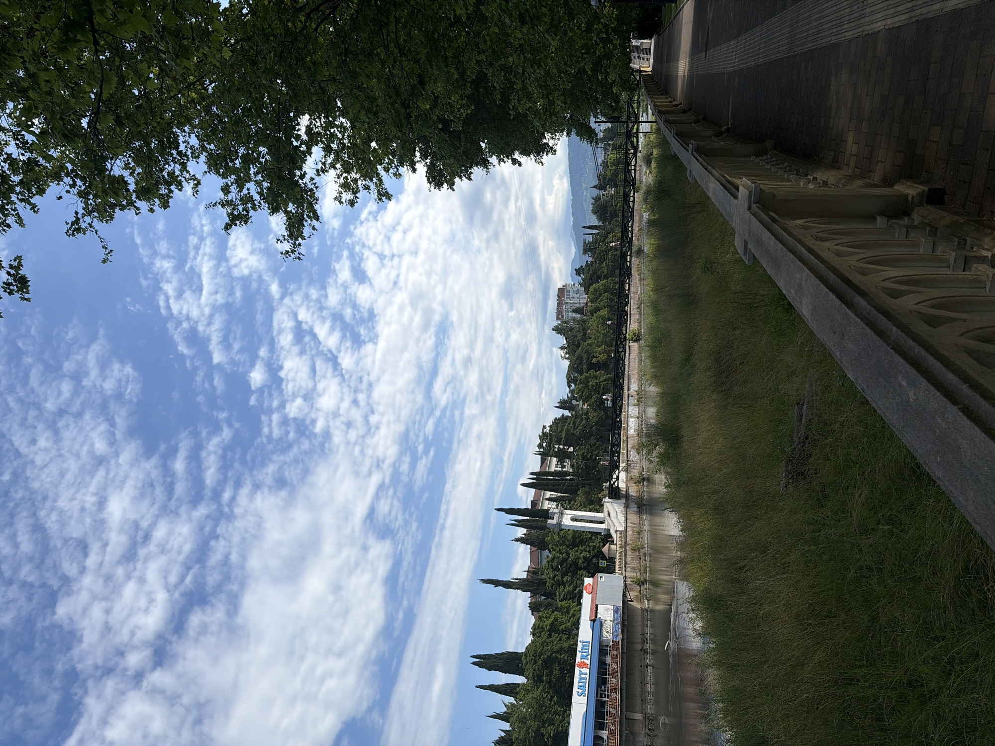 The image captures a sunny day in Sochi, Russia, with a clear blue sky dotted with fluffy white clouds. In the foreground, a paved walkway runs alongside a decorative stone balustrade, bordered by a lush green lawn. The walkway leads towards a bridge crossing a wide river. On the opposite bank, a building with a blue facade and the text "SANIT Rini" is visible, nestled among tall cypress trees and lush greenery. Further in the distance, buildings and mountains can be seen under the expansive sky. The scene is serene and picturesque, showcasing a pleasant urban landscape.