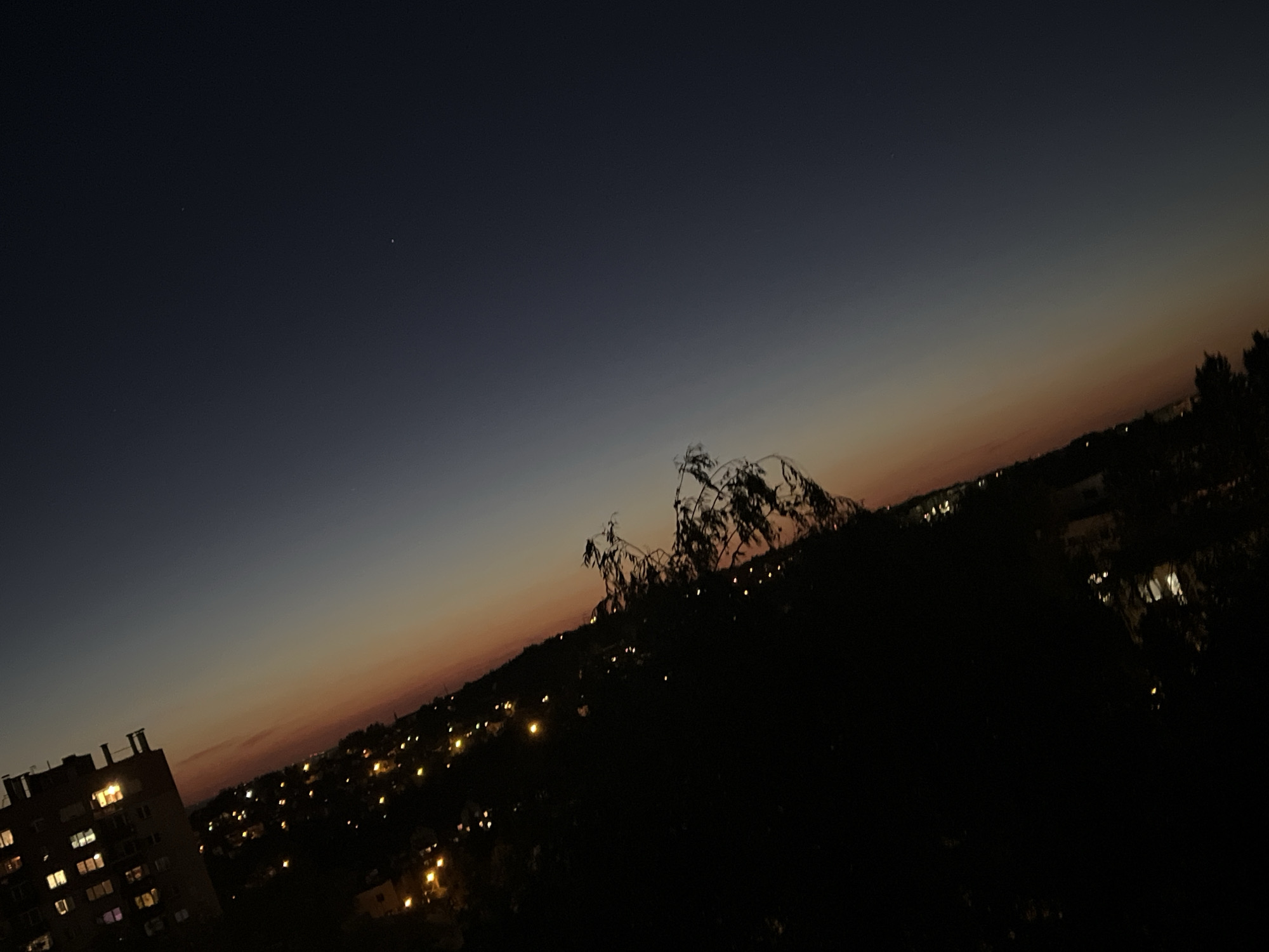 A nighttime cityscape under a dark blue and orange gradient sky. In the lower left is a lit apartment building. Scattered lights indicate houses on a hillside. A dark mass of trees and foliage occupies the central part of the image. The location is Unknown, Unknown.