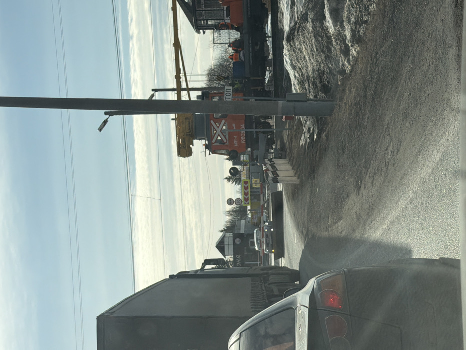 The scene depicts a daytime view of a road approaching a railway level crossing in 29-Y Km, Russia.
In the immediate foreground, the rear of a dark-colored sedan, possibly a Mitsubishi Lancer, is visible on the left, with its left taillight illuminated. To its right, a large dark semi-truck is stopped. Further ahead, a white car with headlights on is stopped at the level crossing, facing the camera. The road surface is asphalt mixed with gravel and dirt, and partially covered in melting snow and slush on the shoulders.
On the right side of the image, a red railway track machine, identified by "МПТ-4 ЖЕЛДОР 1980834" and a white 'X' symbol, is positioned on the tracks. It features an elevated yellow crane-like mechanism. Adjacent to the machine, a white sign with "СТОП" (STOP) in red letters is visible. Multiple individuals, wearing orange high-visibility vests, are present near the railway machine and on an associated platform, engaged in apparent work activities.
The level crossing is marked by yellow and black striped barriers or temporary construction barriers. In the background, a small control building or guardhouse is visible, along with coniferous trees. Overhead utility lines traverse the bright blue sky, which contains scattered, thin white clouds. A prominent utility pole with a street lamp stands on the right side of the road. Signs indicating a speed limit of "30" are also visible.