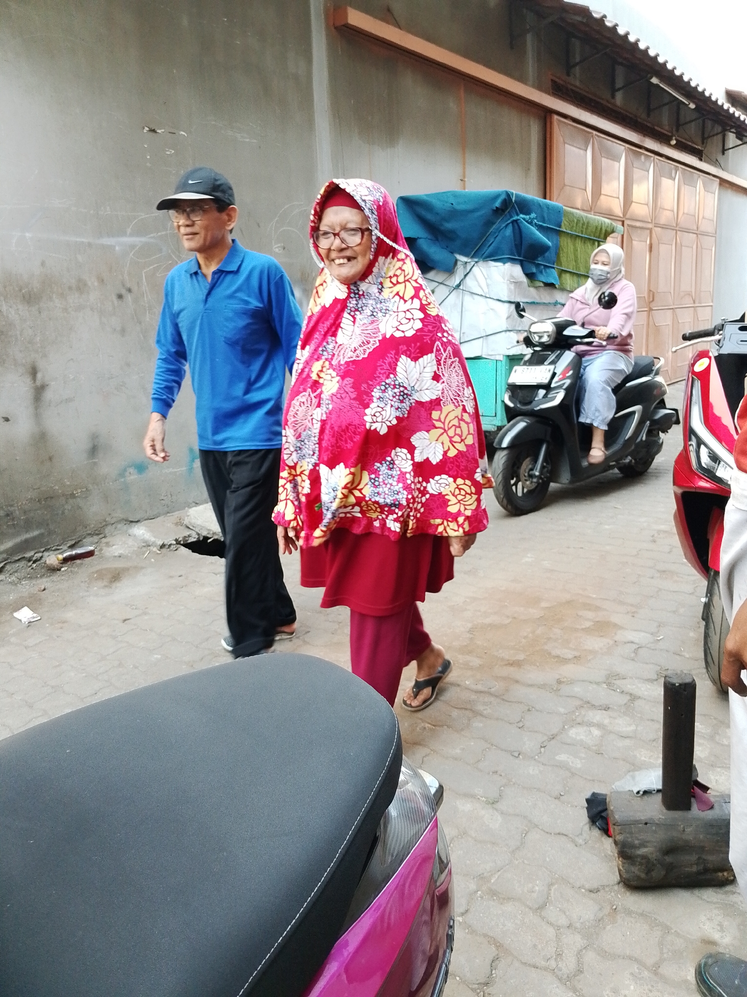 In a city street in Pasuruan, Indonesia, a man wearing a blue long-sleeved shirt, black cap, and glasses walks to the left. Beside him, a woman in a bright red hijab with a floral pattern and a red skirt smiles. Behind them, a woman on a black scooter wears a light pink long-sleeved shirt, light blue pants, and a light gray hijab and mask. In the foreground, the seat of a dark gray scooter is visible. The environment includes a textured concrete ground, a light gray wall, and a building with a metal roof and brown paneled doors.