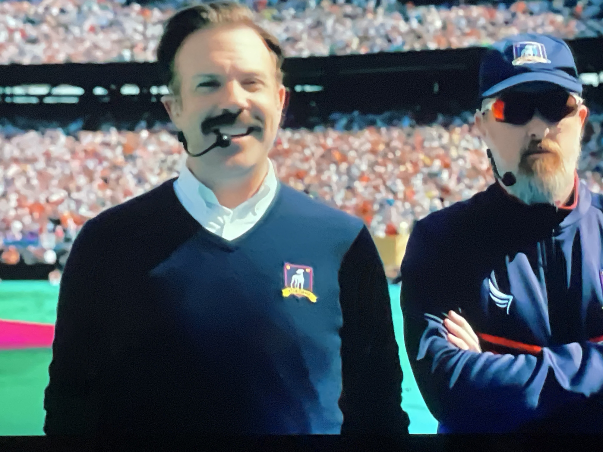 Two men are standing in front of a blurred crowd of spectators in a stadium. The man on the left is wearing a dark V-neck sweater over a white collared shirt, with a microphone attached to his ear. He has a mustache and is smiling. On his sweater, there is a crest with a dog inside a shield. The man on the right is wearing a dark blue jacket and a blue baseball cap with a similar crest on it. He has sunglasses on and a goatee. His arms are crossed. The environment suggests a sporting event.