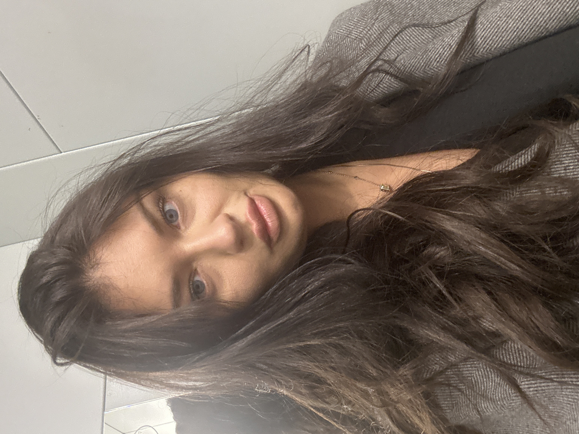 A close-up portrait-style image, rotated 90 degrees counter-clockwise, features a young woman looking directly at the camera. She has long, dark brown hair that cascades around her face and shoulders, appearing somewhat dishevelled. Her large, blue-grey eyes are prominent, and her lips are slightly parted in a subtle pout. She is wearing a delicate golden necklace with a small, light-colored pendant. Her attire includes a grey jacket or coat with a clear herringbone pattern, worn over what appears to be a darker top. The background is a plain, light-colored surface, possibly a wall with faint lines suggesting tiles or panels. An indistinct reflection can be seen in the lower left corner. The scene is indoors, and no specific time of day or weather is indicated. There is no visible text. The woman's expression is neutral yet engaging.