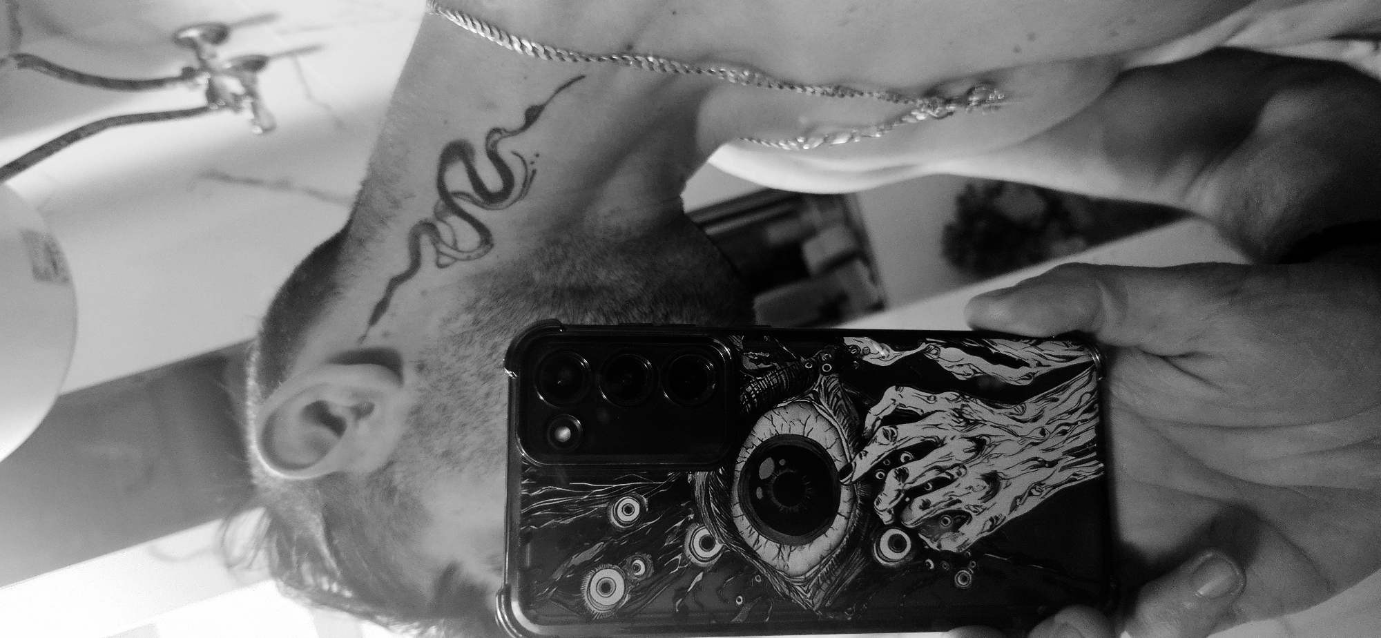 A person takes a selfie in what appears to be a bathroom mirror. The person has a snake tattoo on their neck and is wearing a silver chain. They are holding a smartphone with a black case featuring a stylized eye and hand design. The reflection shows part of the bathroom, including the sink fixtures. The location in Plovdiv, Bulgaria is not determinable from the image.