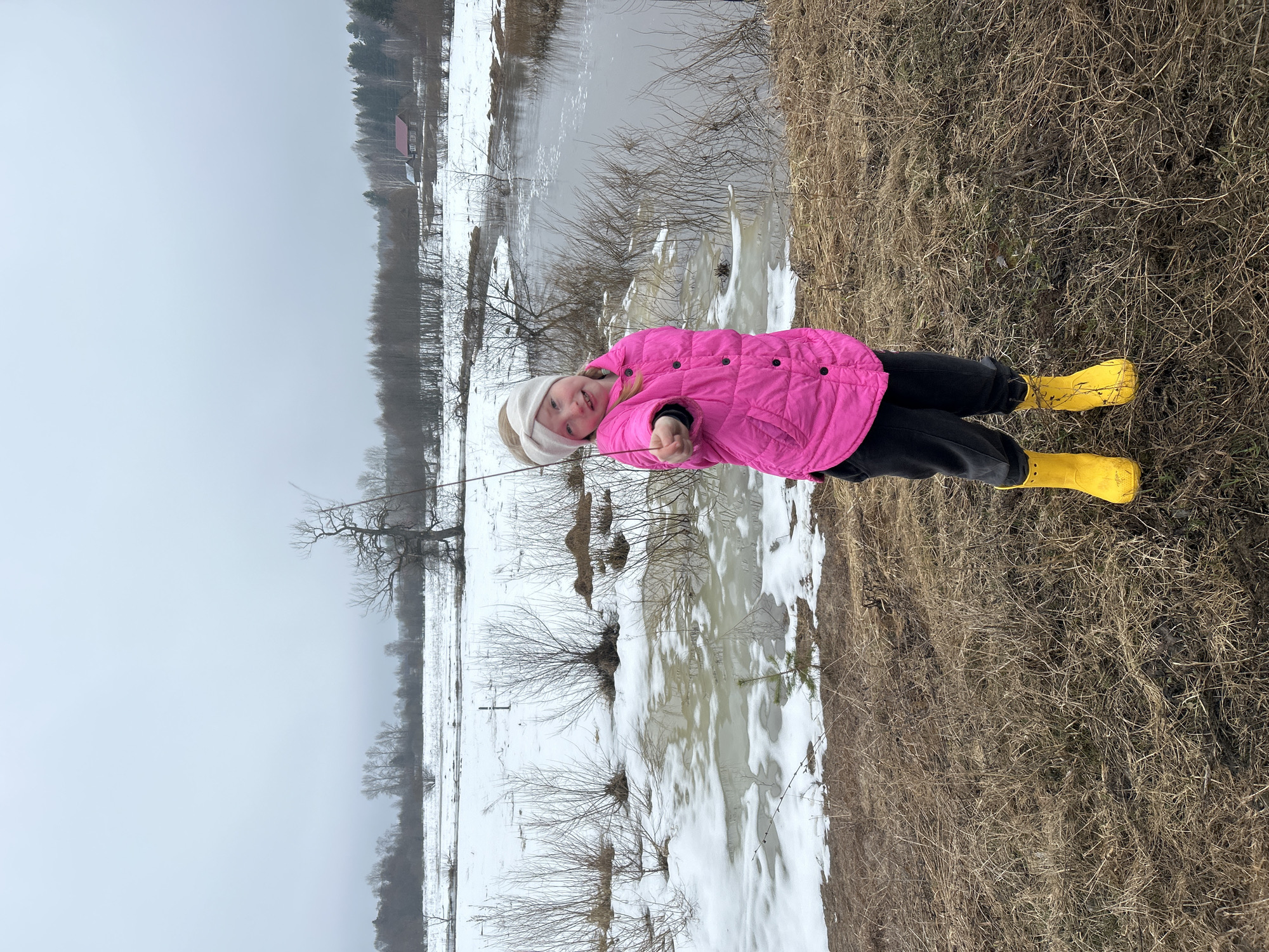 A girl stands in a field in Уральское, Russia. She wears a pink jacket, black pants, a white hat, and yellow boots. The background shows a field with snow and water, with bare trees and a building in the distance under a gray sky. The ground near her is covered in dry grass and snow patches. She is holding a thin stick in her hands.