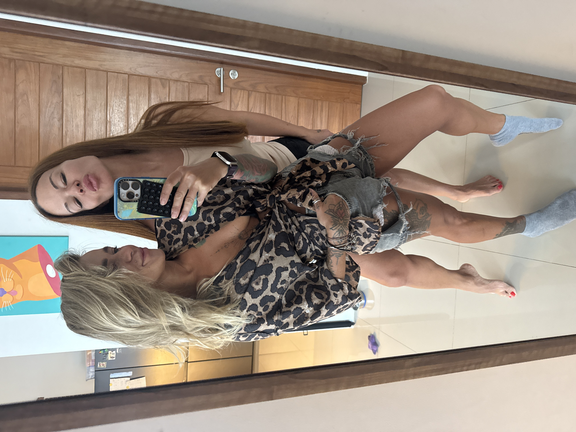 Two women are taking a mirror selfie in what appears to be a residential interior. The woman in the foreground has blonde hair and is wearing a leopard-print shirt tied at the waist, ripped denim shorts, and is holding a phone. She has tattoos on her arms and chest. The woman behind her has long brown hair and is wearing a beige tank top and black shorts, puckering her lips for the photo. She is also holding the phone. The background shows a wooden door, a refrigerator, and a colorful painting of a cat on the wall. The floor is tiled.