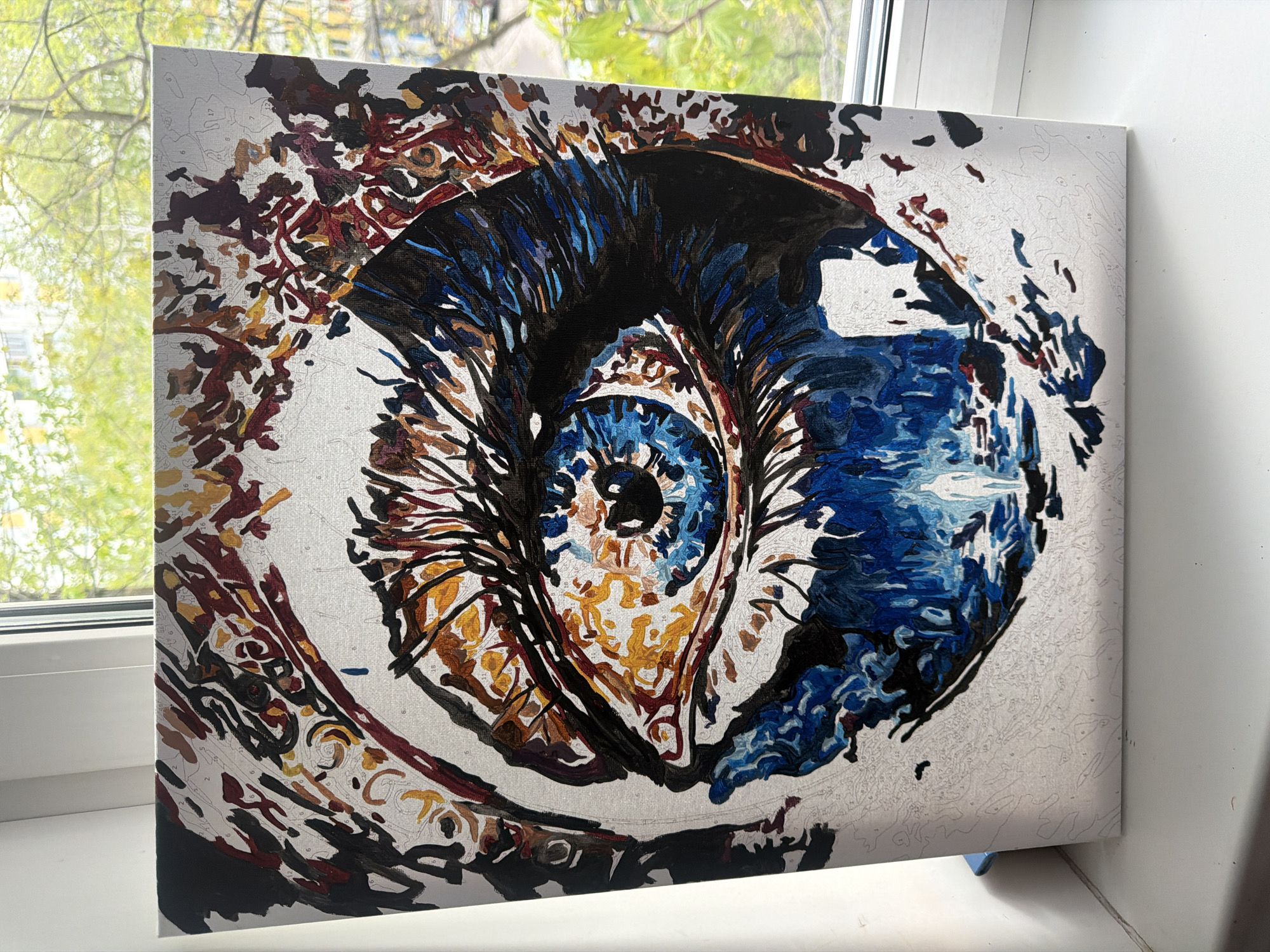 The image shows a close-up of a partially completed paint-by-numbers artwork depicting an eye. The artwork is on a canvas resting on a white windowsill. Outside the window, blurred green foliage suggests a tree. The painting's iris is predominantly blue, with the surrounding area featuring black, brown, gold, and white. Unpainted sections of the canvas reveal numbered areas yet to be filled in. The location is not identifiable as Moscow, Russia, based on visual information.