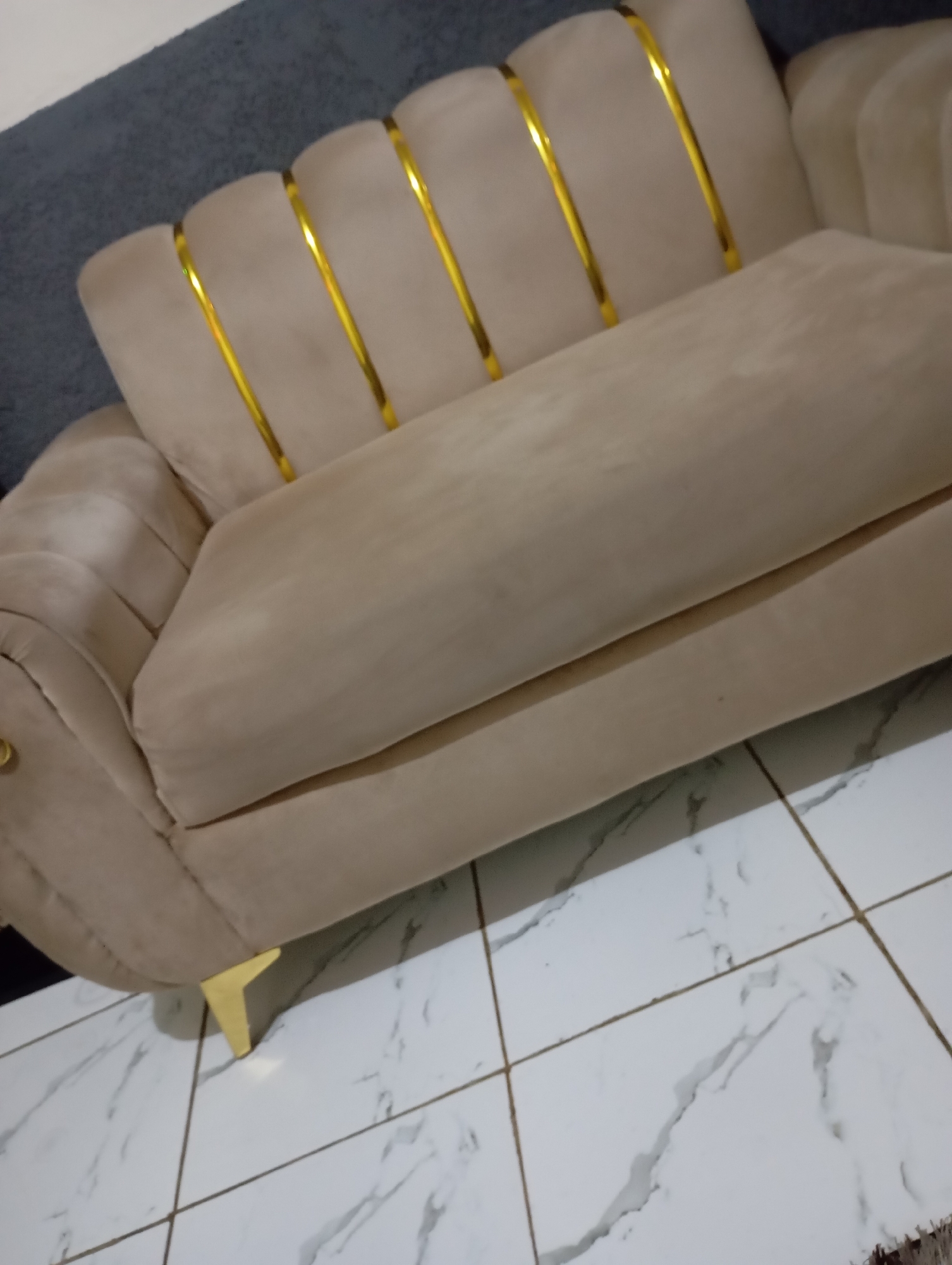 The image shows a close-up, slightly angled view of a light beige, plush sofa with gold decorative accents. The sofa features channel tufting on its backrest, with vertical gold bands accentuating the channels. A large seat cushion is visible, extending forward. The sofa has golden, tapered legs. The floor beneath the sofa is tiled with a white marble pattern and dark grout lines. The background shows a dark gray upholstered surface, possibly another piece of furniture or a wall covering, and a sliver of white. The scene is indoors.
