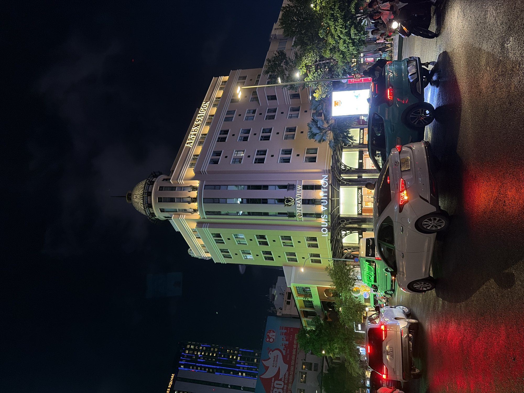 The image presents a night view of a city street in Thủ Đức, Vietnam. The ARTEX Saigon building stands prominently, with the signs "Opera View" and "Louis Vuitton" visible on the building's storefront. Street lighting illuminates the scene. Several vehicles, including cars and motorbikes, are in the street, suggesting traffic. A pedestrian crowd can be observed on the sidewalk adjacent to the street. Advertisements are visible on other buildings in the background.