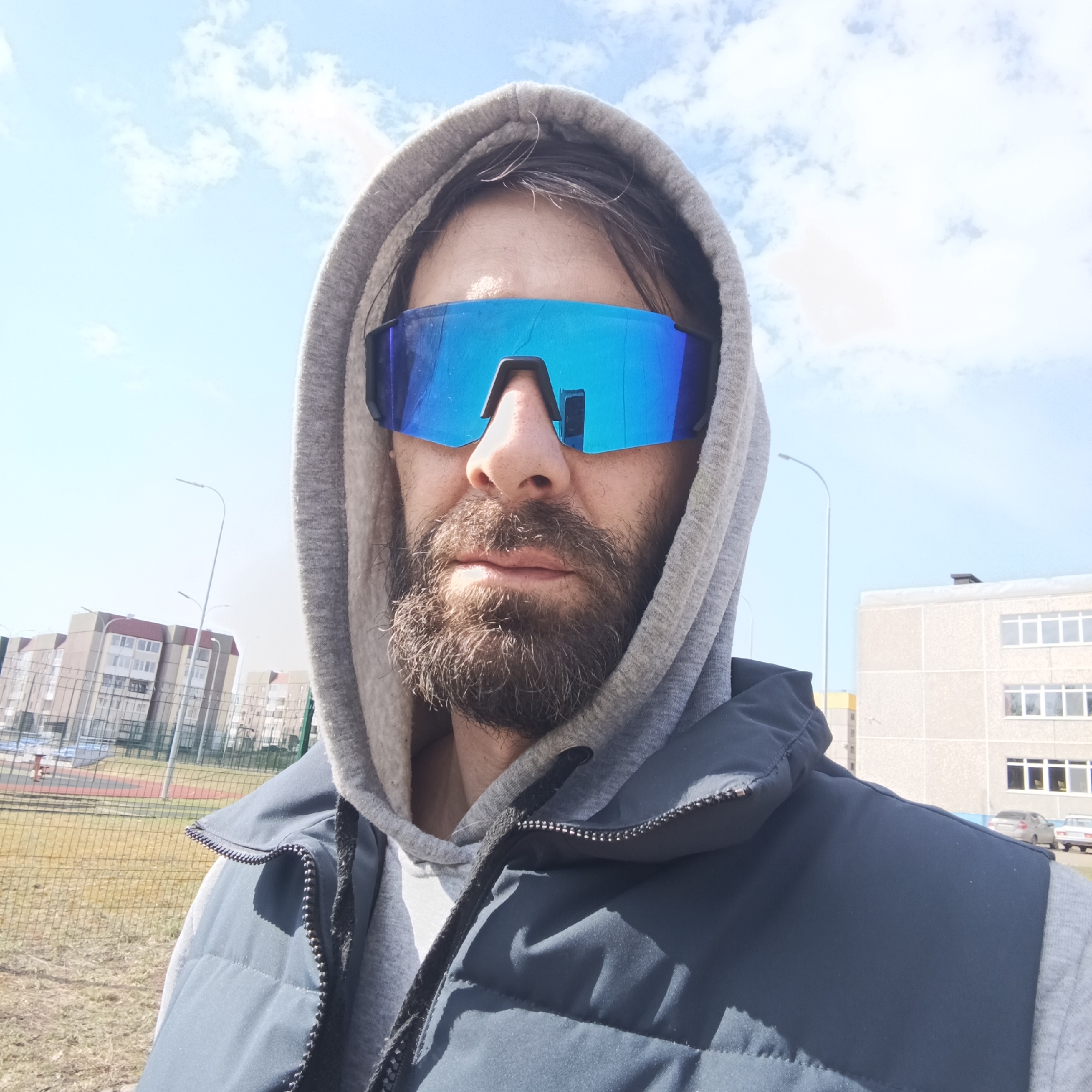 The image is a medium shot of a man with a beard, wearing a gray hoodie and a navy blue vest. He is also wearing reflective blue sunglasses. The location appears to be in an urban area, possibly Markovskii, Russia, based on the architecture. In the background, there are apartment buildings and a fenced-off playground. The sky is visible, with some clouds. The time of day seems to be daytime, based on the natural light.