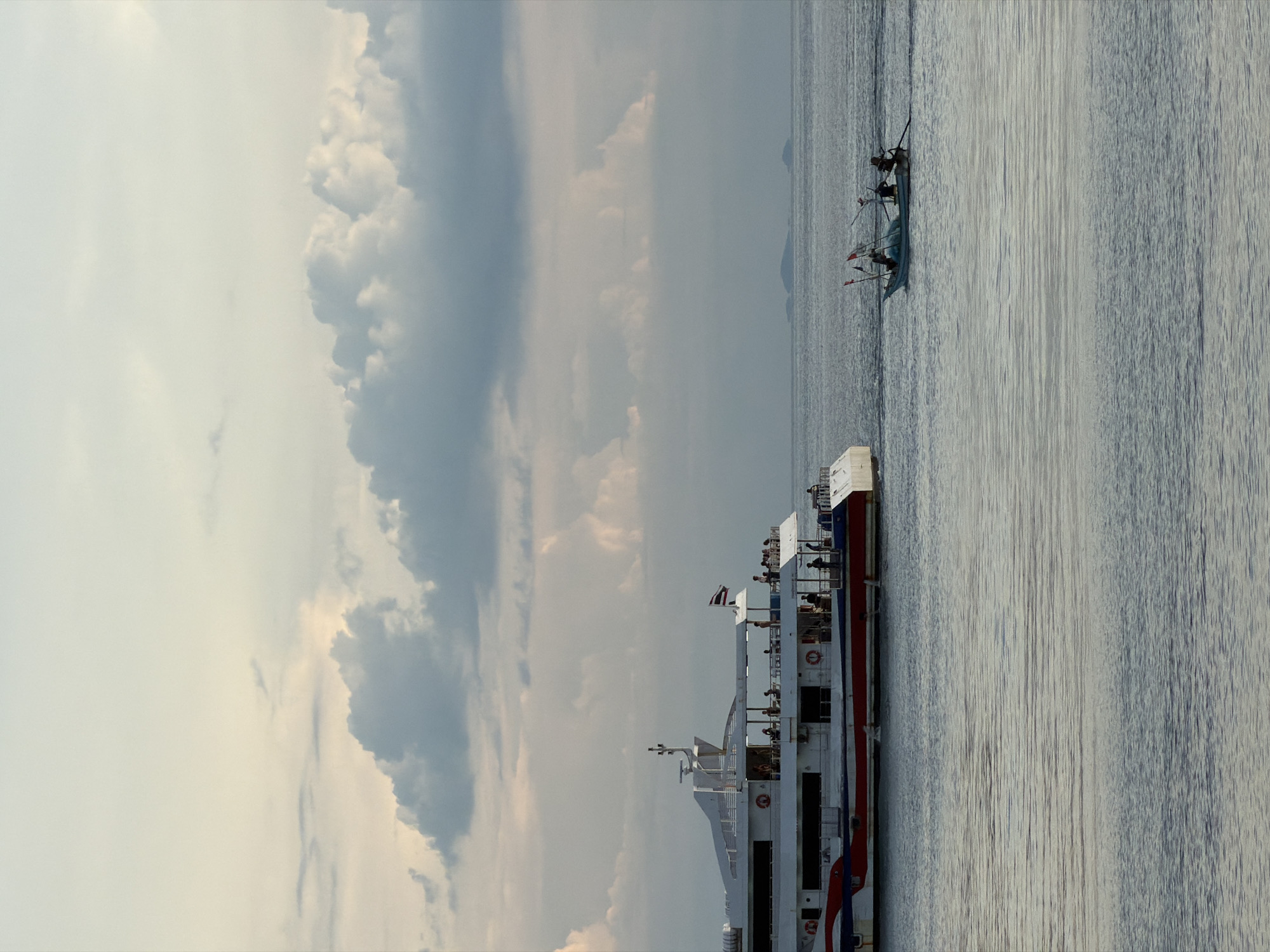 A ferry boat carrying passengers navigates the calm sea off the coast of Koh Phangan, Thailand. The ferry, a long white vessel with red and blue accents, has multiple decks where people can be seen standing. Life preservers are visible on the side of the ferry. In the distance, a smaller fishing boat with three people on board is also visible. The sea is a light blue-gray, reflecting the cloudy sky above. The sky is a mix of light and dark clouds, suggesting it might be late afternoon or early evening. The overall atmosphere is peaceful, with the boats moving smoothly through the water under a dramatic sky. There is a small flag with a red and white pattern visible on the ferry.