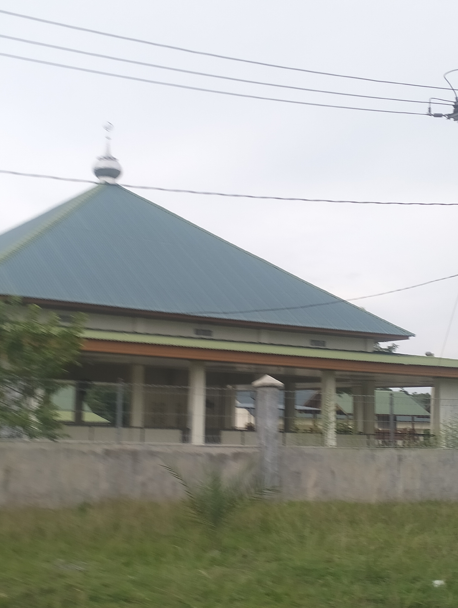 This image shows a mosque in Lhokseumawe, Indonesia. The mosque has a prominent, steeply pitched teal-colored metal roof, topped with a small white dome and crescent moon finial. A light-colored building with a green roof and orange trim extends from the main structure, supported by visible pillars. A concrete wall runs across the foreground, partially obscuring a fenced area with additional buildings or structures visible beyond. Overcast sky and power lines are visible above the mosque. The foreground is filled with blurred green grass and vegetation, suggesting the photo was taken from a moving vehicle. There are no people visible in the image.