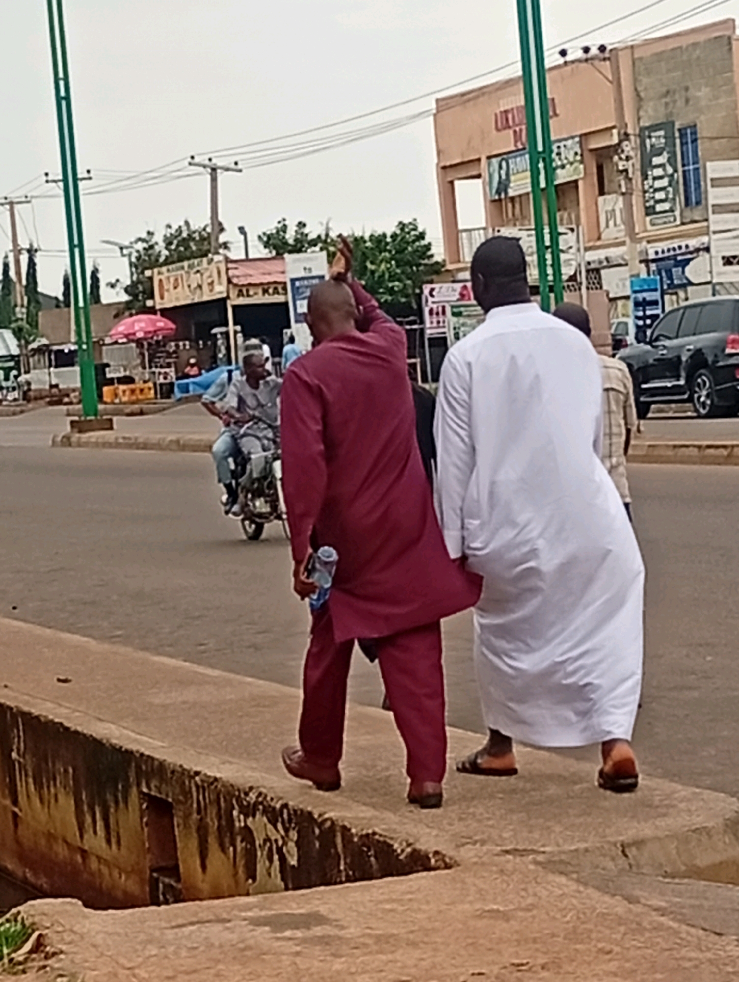Two men are walking away from the camera along a raised concrete walkway. The man on the left is wearing a maroon kaftan and brown shoes, and is holding a clear plastic bottle in his right hand. The man on the right is wearing a long white robe and brown sandals. They are walking next to a street where a motorcycle is being ridden by another man in a light blue shirt and jeans. In the background, there is a row of buildings, including one with "AL-KAS" visible on a sign. There are also utility poles with wires. The scene appears to be in an urban area, likely a marketplace or commercial street, possibly in Garko, Nigeria, given the architectural style and clothing. The sky is overcast.