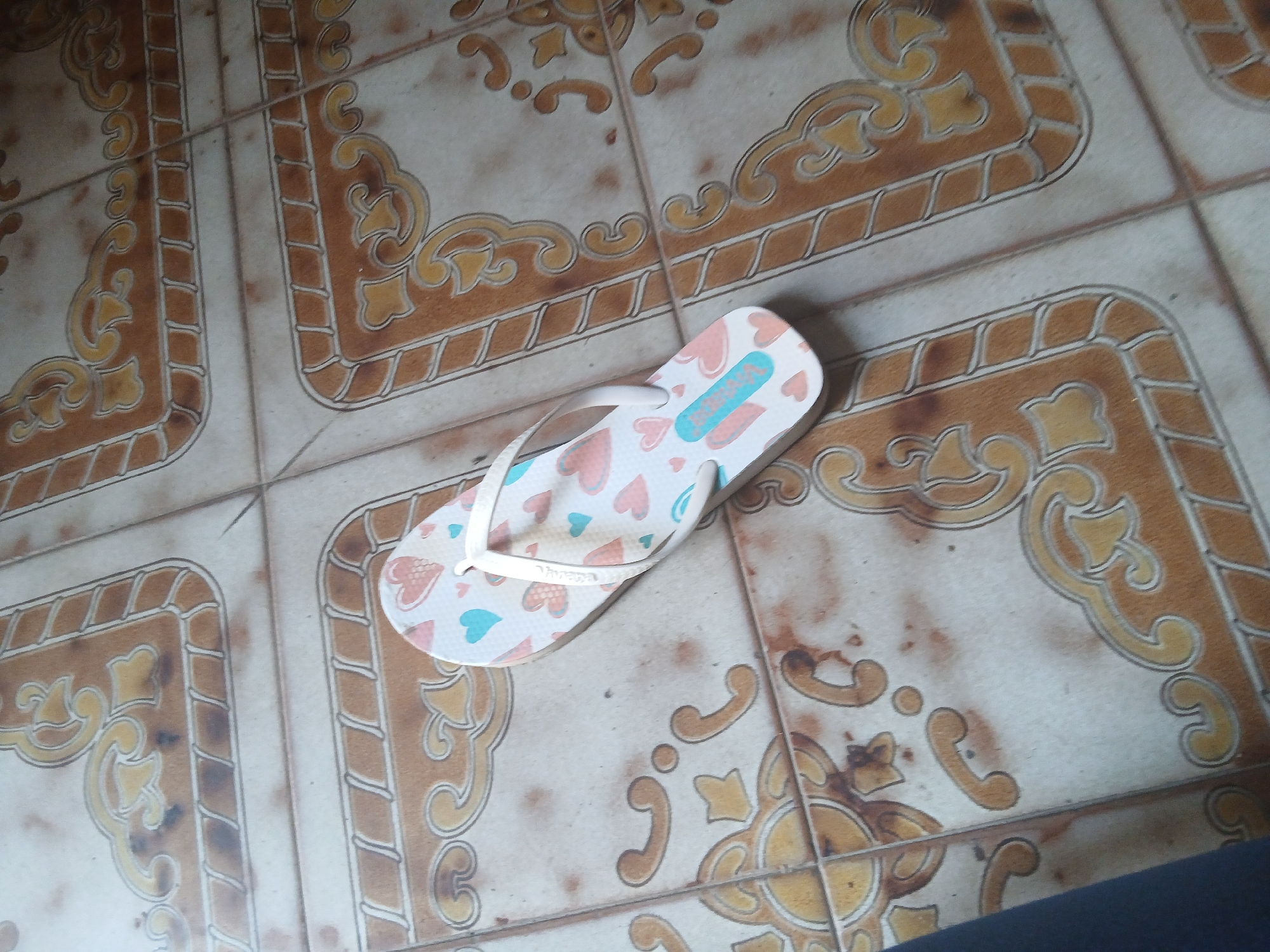 A single white flip-flop with a heart pattern is lying on a tiled floor. The tiles have a decorative pattern with brown and yellow elements. The flip-flop is white with a pattern of pink and light blue hearts. The straps of the flip-flop are white. The image appears to be taken indoors, possibly in a residential setting. The location of Santa Ana, Venezuela is not discernible from the image itself.