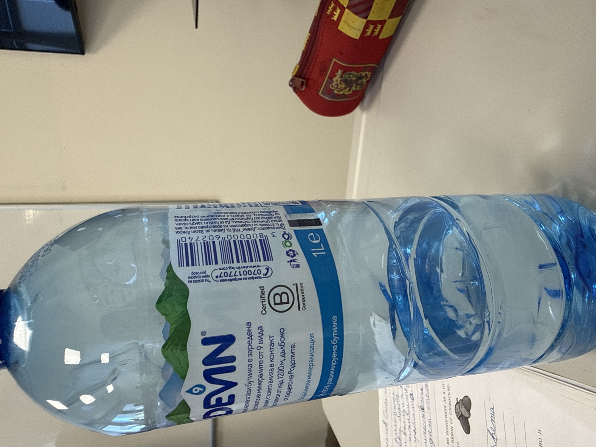 Here's a description of the image:
**Objects:** The primary object is a plastic bottle of Devin brand mineral water, appearing mostly full. A red pencil case is partially visible near the top. A sheet of paper with handwriting and a small drawing (possibly of a mushroom) sits beneath the water bottle.
**Environment:** The objects rest on a light-colored surface. The background is minimal, with parts of other unidentifiable objects (a black rectangular object and another item on the right edge of the frame) showing out of focus.
**Label Details:** The label on the bottle includes the brand name "DEVIN," information about the water's mineral content, certification logos, recycling information, and contact details for the consumer. The label is in Bulgarian. The bottle indicates it contains 1 liter of water.