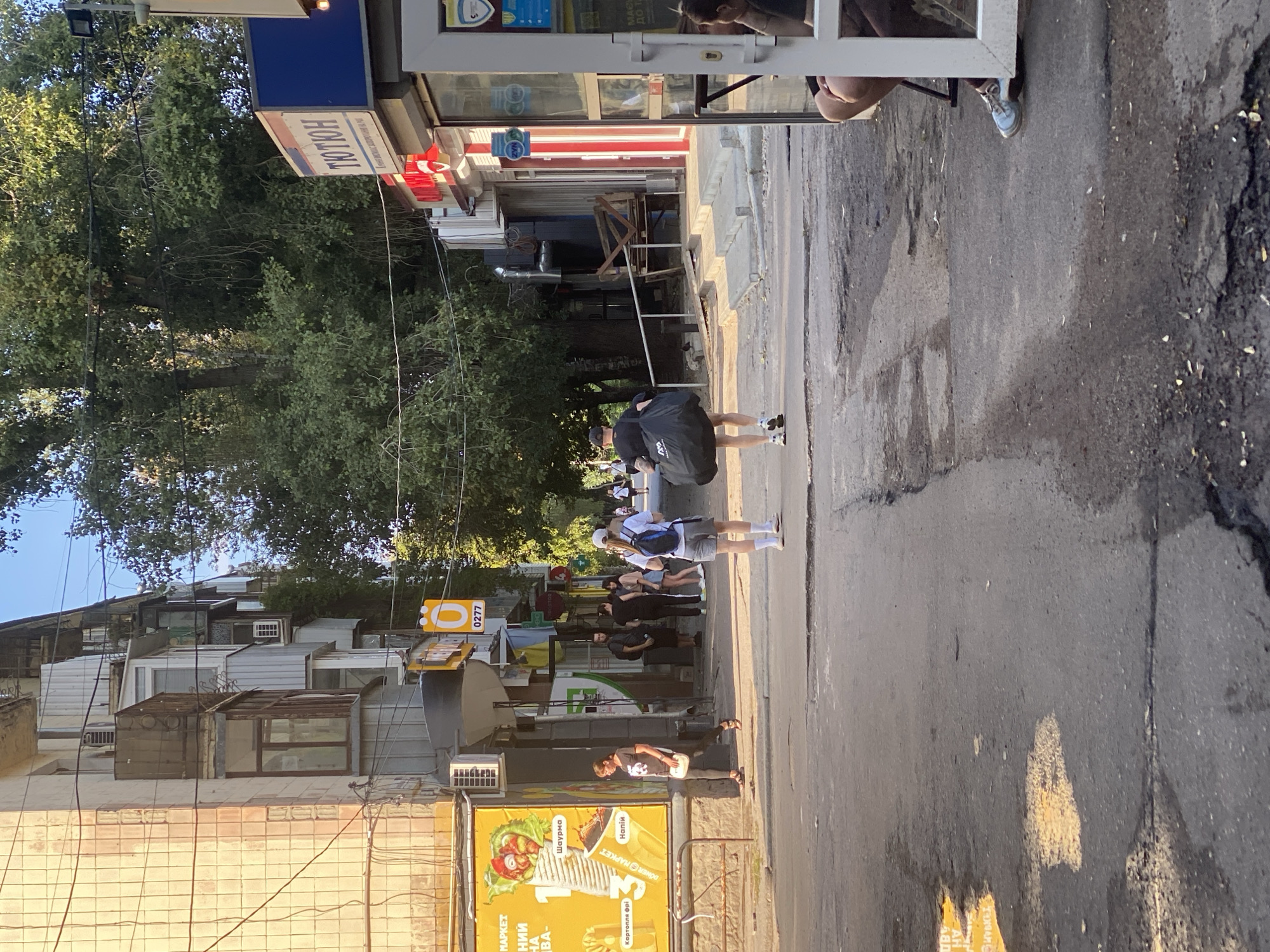 The image depicts a street scene in Poltava, Ukraine, during daylight hours. The perspective is from a ground-level view, looking down a paved street lined with buildings on the left and a row of shops or kiosks on the right. Tall trees with dense foliage are visible in the background, creating a canopy over the street in places.
On the left side of the street, a building with a beige, tiled facade is prominent. It features multiple balconies and windows. A large yellow advertisement poster with images of food and text is affixed to the side of this building.
On the right side of the street, a series of shops or kiosks with awnings and signs are present. One sign reads "ТЮТЮН" in large letters. Another appears to be a food vendor with red and white striped accents. A white-framed structure, possibly a kiosk with glass panels, is in the foreground on the right, revealing a glimpse of a person seated inside.
Several people are visible walking along the street. In the mid-ground, a person is walking away from the camera, carrying a large black duffel bag over their shoulder. This person is wearing a black t-shirt, shorts, and a baseball cap. Further ahead, two individuals are walking towards the camera. One is wearing a white t-shirt, grey shorts, and a white cap, with a blue backpack. The other is wearing a light-colored t-shirt and dark shorts. A woman in a black t-shirt and dark pants is walking on the left side of the street towards the background, carrying a white purse. Other individuals are scattered in the background, appearing to be pedestrians.
The street surface is asphalt and shows signs of wear, with cracks and some unevenness. Puddles of water and debris are also visible on the pavement. The overall impression is of a typical urban street in a residential or commercial area.