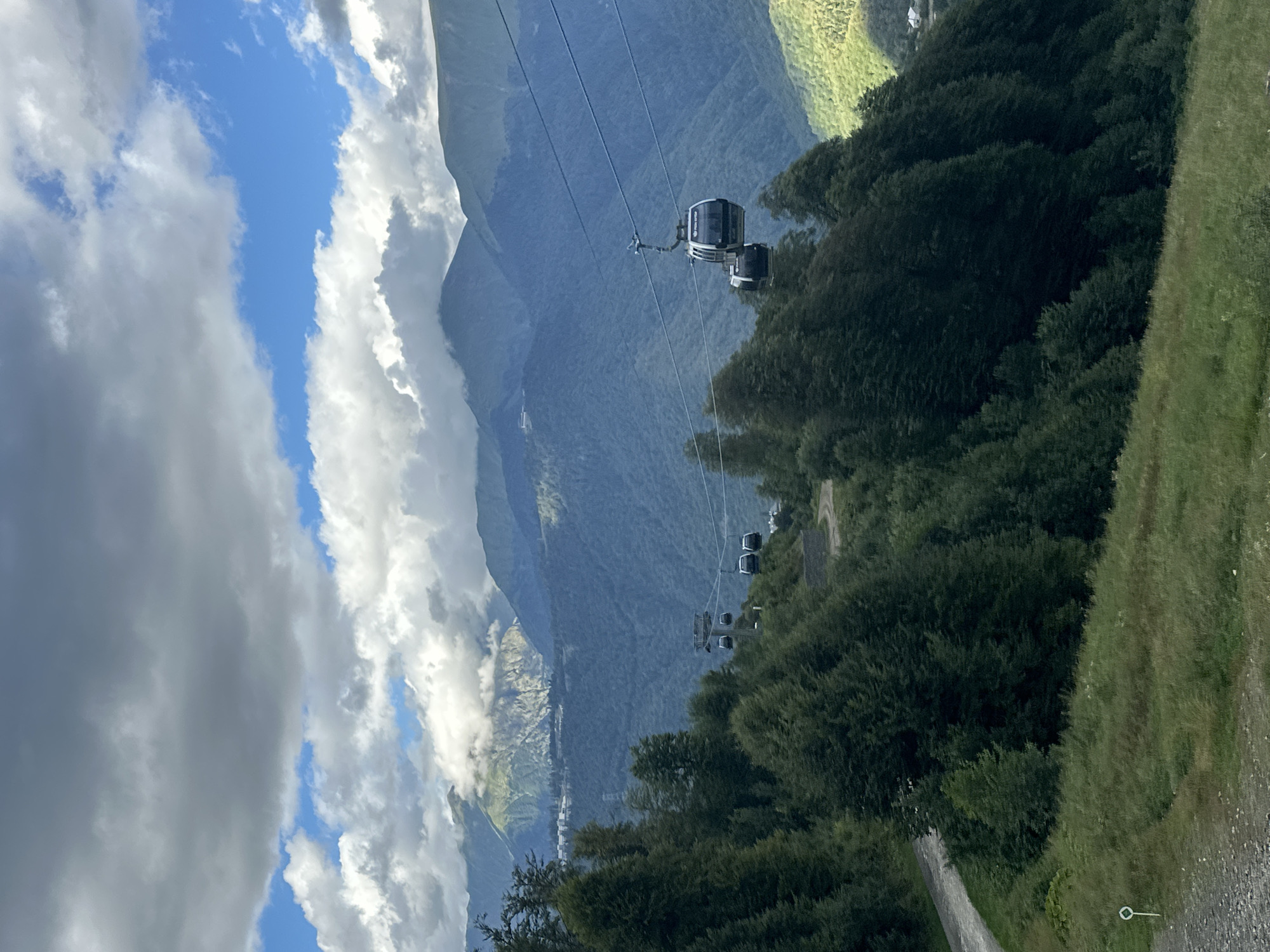 The image depicts a scenic mountain landscape in Estosadok, Russia, characterized by lush green slopes, towering trees, and a dramatic sky filled with clouds. In the foreground, a winding dirt path cuts through grassy terrain. The middle ground is dominated by dense evergreen forests that cascade down the mountainside. A cable car system is prominently featured, with several cabins suspended from cables, transporting people between different elevations. The cable car cabins are modern and sleek, with large windows offering panoramic views of the surrounding scenery. In the background, majestic mountain ranges rise, their peaks partially obscured by swirling clouds. The overall impression is one of natural beauty and outdoor adventure, with the cable car serving as a vital mode of transportation for exploring the mountainous terrain. The atmosphere is serene and tranquil, with the play of light and shadow adding depth and dimension to the scene.