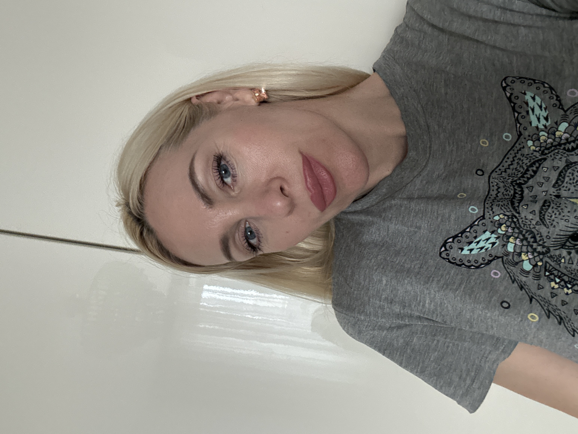 This media file is a close-up, head-and-shoulders portrait of a young Caucasian woman with blonde hair. She is wearing a grey t-shirt with a graphic print of an owl on it. The image is oriented sideways, with the woman's head tilted. She has blue eyes, visible makeup including mascara and lipstick, and a small rose-gold earring in her visible ear. The background appears to be a plain, light-colored wall. The lighting suggests an indoor setting, possibly during the day. Her expression is neutral, with a slight tilt of her head. No text is visible in the image.