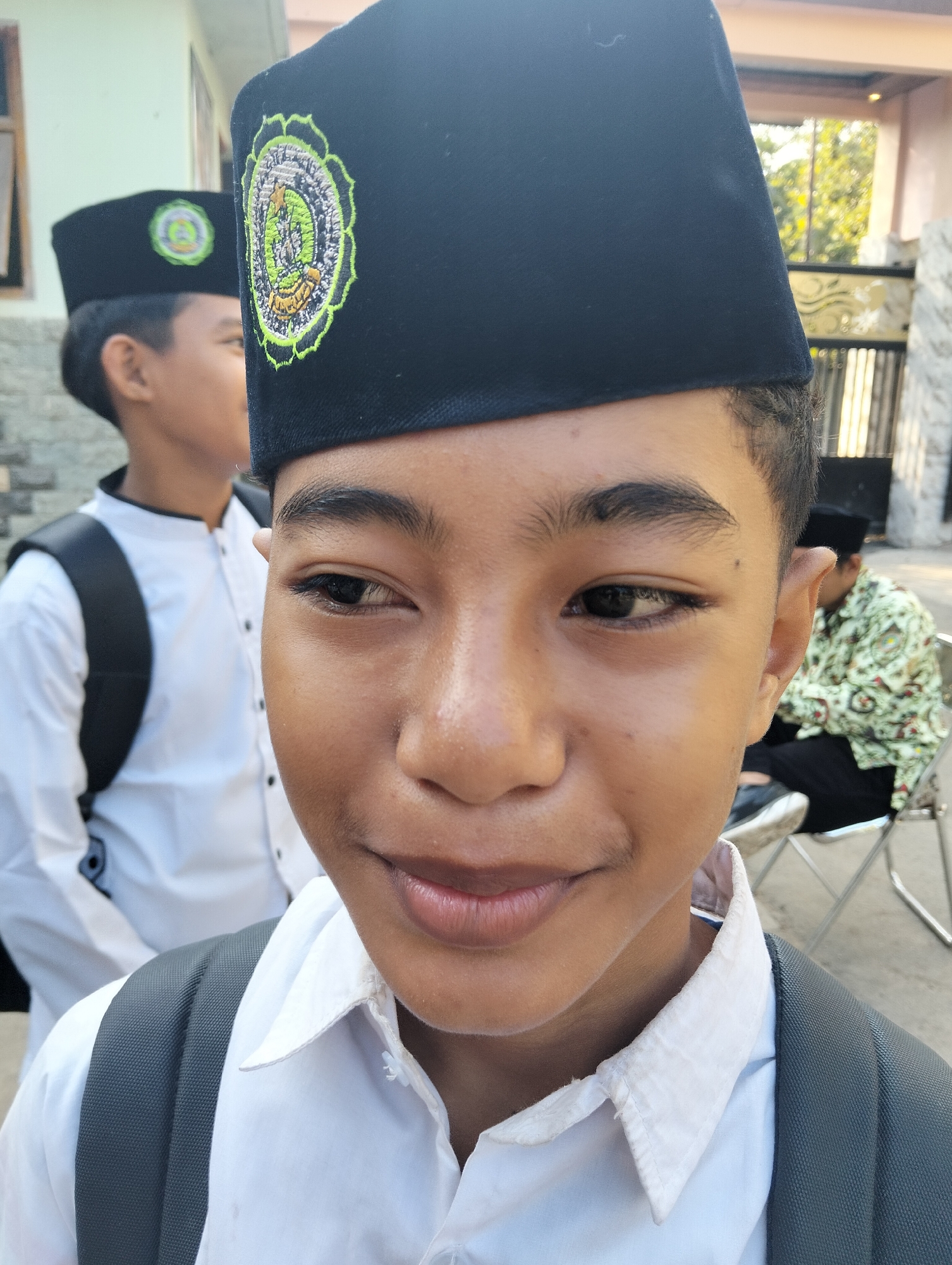 A young boy, wearing a black peci hat with an embroidered green and yellow emblem, smiles as he looks to his left. He is dressed in a white collared shirt and has a grey backpack on. In the background, another boy in a similar peci and white shirt stands out of focus, and an older person sits on a chair wearing a patterned green shirt. The scene appears to be outdoors, possibly in front of a building with a gated entrance and trees visible in the distance. This image was likely taken in Paciran, Indonesia, based on the context of similar images.