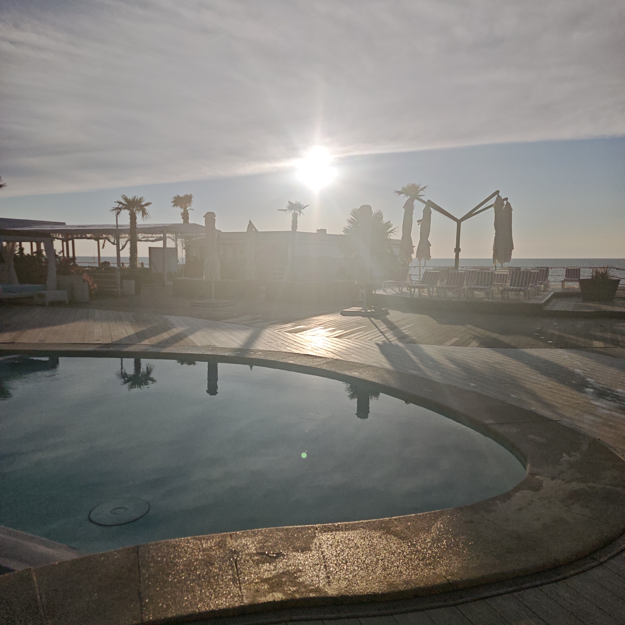 An outdoor swimming pool with a wooden deck is in the foreground. Beyond the pool, there is a beach with lounge chairs and umbrellas, and further in the distance, the ocean is visible under a bright, hazy sun. Several palm trees are scattered throughout the scene, and the sky is a pale blue with wispy clouds. The sun is positioned high in the sky, creating lens flares and casting long shadows. The scene appears to be a resort or a beachside establishment.