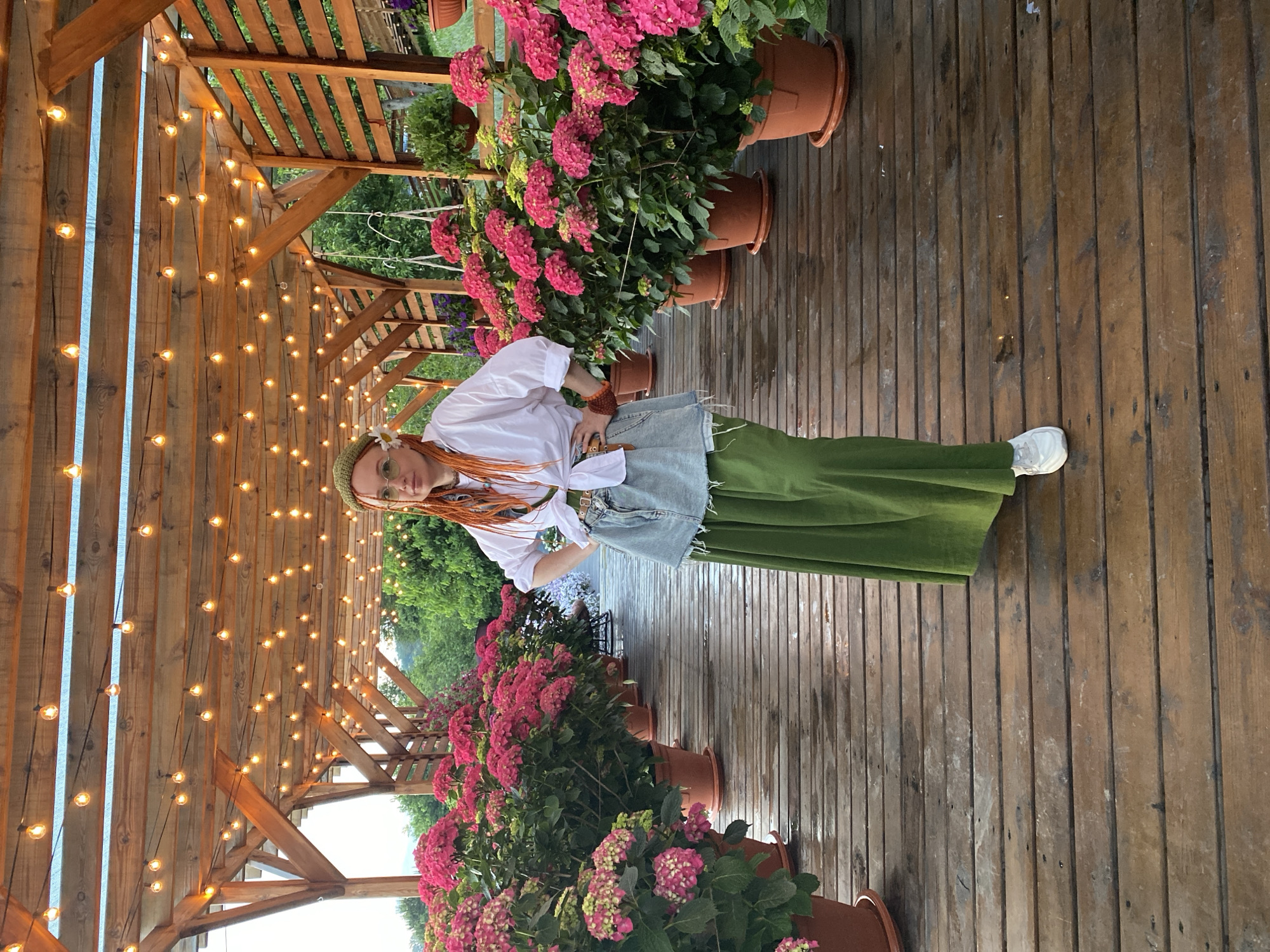 In Verkhneye Uchdere, Russia, a person with vibrant orange hair, wearing a white collared shirt tied at the waist, a denim skirt with frayed edges, and a long green skirt, stands on a wooden deck. The deck is wet, suggesting recent rain. The person is adorned with a green headscarf and a white flower on their left side. They are positioned within a pergola structure, illuminated by strings of warm-toned lights overhead. The pergola is lined with numerous potted hydrangeas featuring abundant pink blooms. Trees and greenery are visible in the background, indicating an outdoor, possibly garden or restaurant setting.