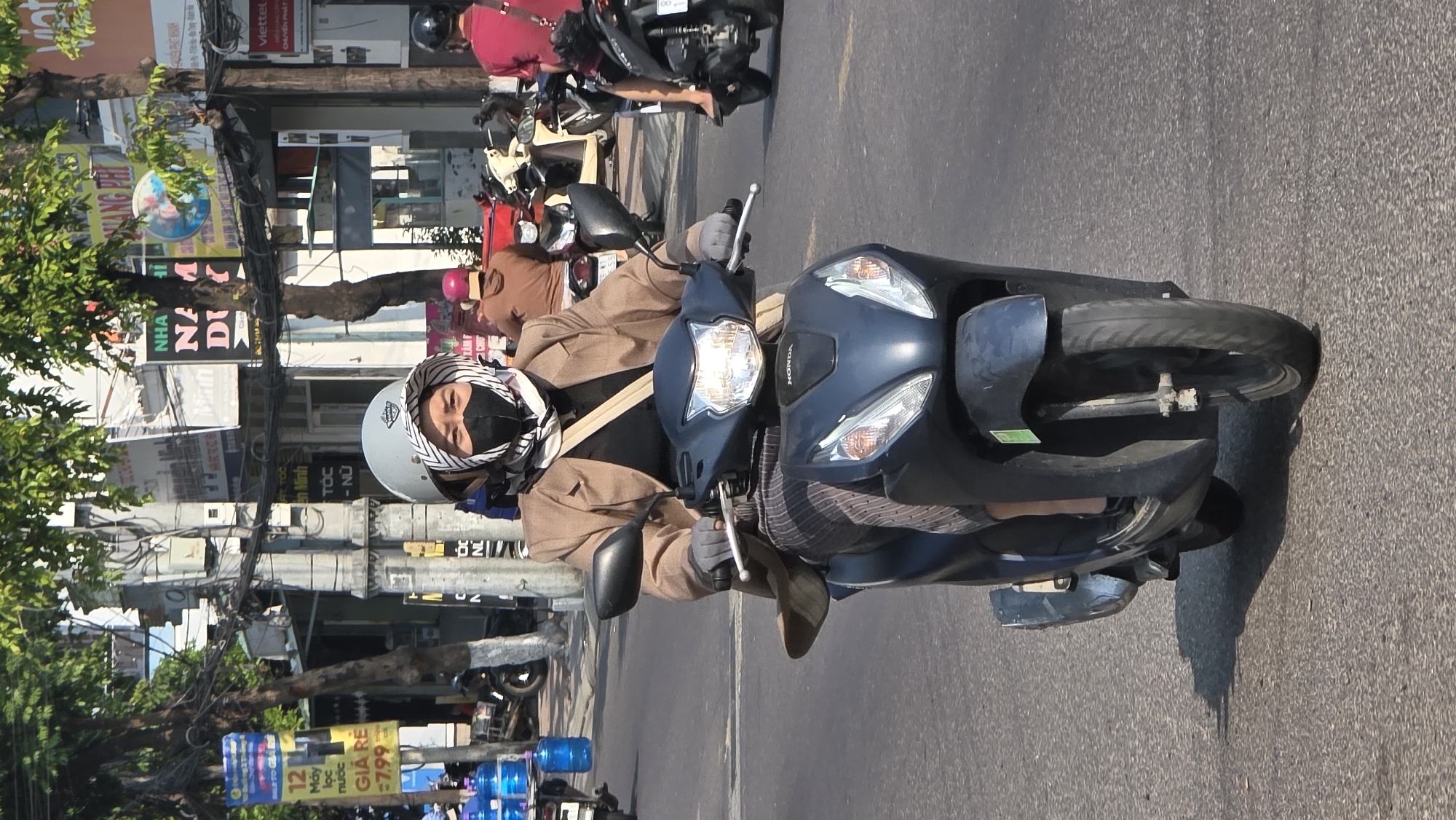The photograph shows a person riding a blue Honda motorcycle on a city street. The rider is wearing a beige helmet with a black and white scarf, a black face mask, a brown jacket, and gray gloves. In the background, there are trees, buildings, and street signs. Signs display the words "Nha Trang" and "Viettel", indicating the location is in the city of Bắc Nha Trang, Vietnam. Other parked motorbikes and people are visible in the background.