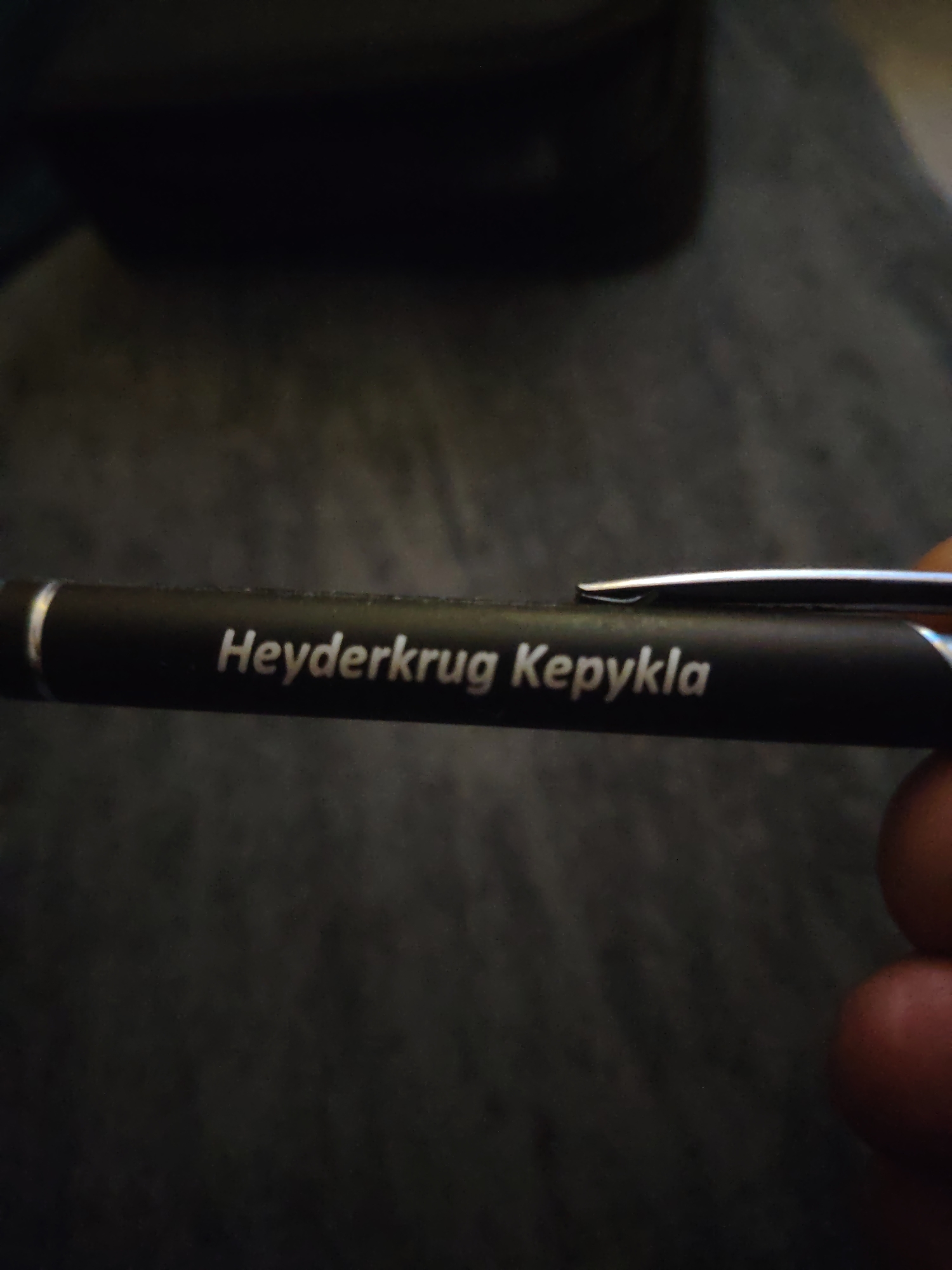 A black cylindrical pen is held horizontally across the center of the frame by a human hand, with the barrel facing the viewer. The pen features a silver-colored clip extending from its top right side and a thin silver band near its left end. Engraved or printed in white capital letters on the pen's black surface is the text "Heyderkrug Kepykla". A thumb and part of an index finger, consistent with a right hand, are visible on the right side of the pen, securing its position. The background is a dark, out-of-focus surface, possibly a textured fabric or desk, with a hint of a darker, undefined object in the upper left background. The lighting highlights the white text on the pen. No specific geographical identifiers for Huntingdonshire, United Kingdom, are visible within the image.
