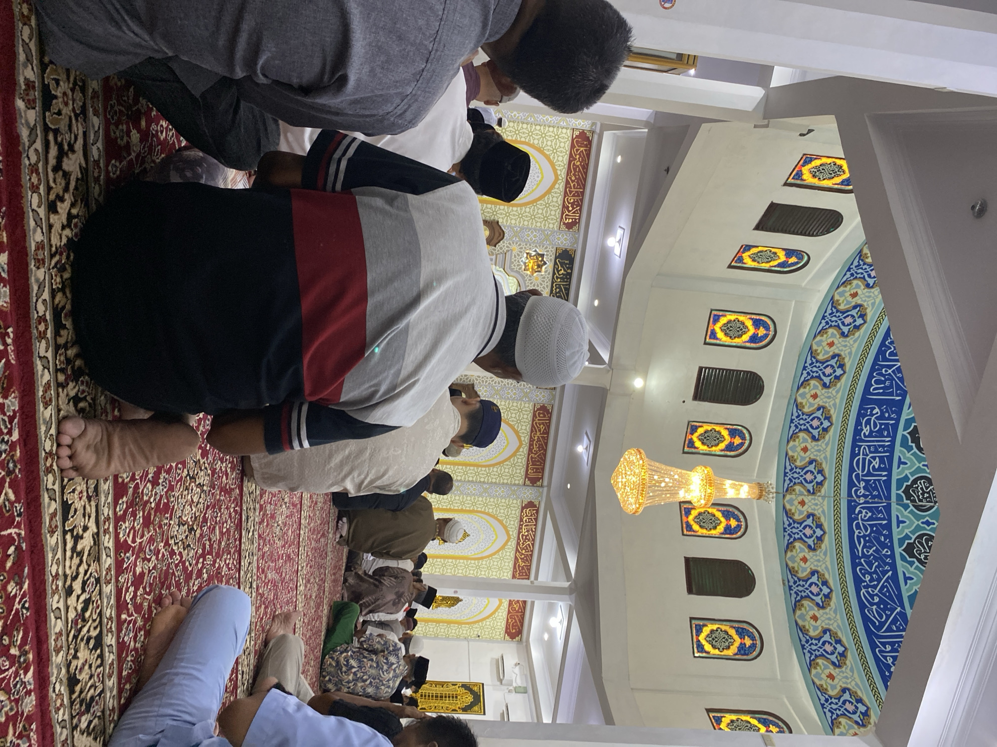The image depicts the interior of a mosque, likely in Lhokseumawe, Indonesia, where a group of men are engaged in prayer. They are seated on a red carpet with intricate patterns. The mosque features high ceilings adorned with colorful Islamic calligraphy and ornate stained-glass windows. A chandelier hangs from the center of the dome. The men are wearing traditional Islamic attire, including skullcaps and long shirts. The scene conveys a sense of spiritual devotion and community gathering.