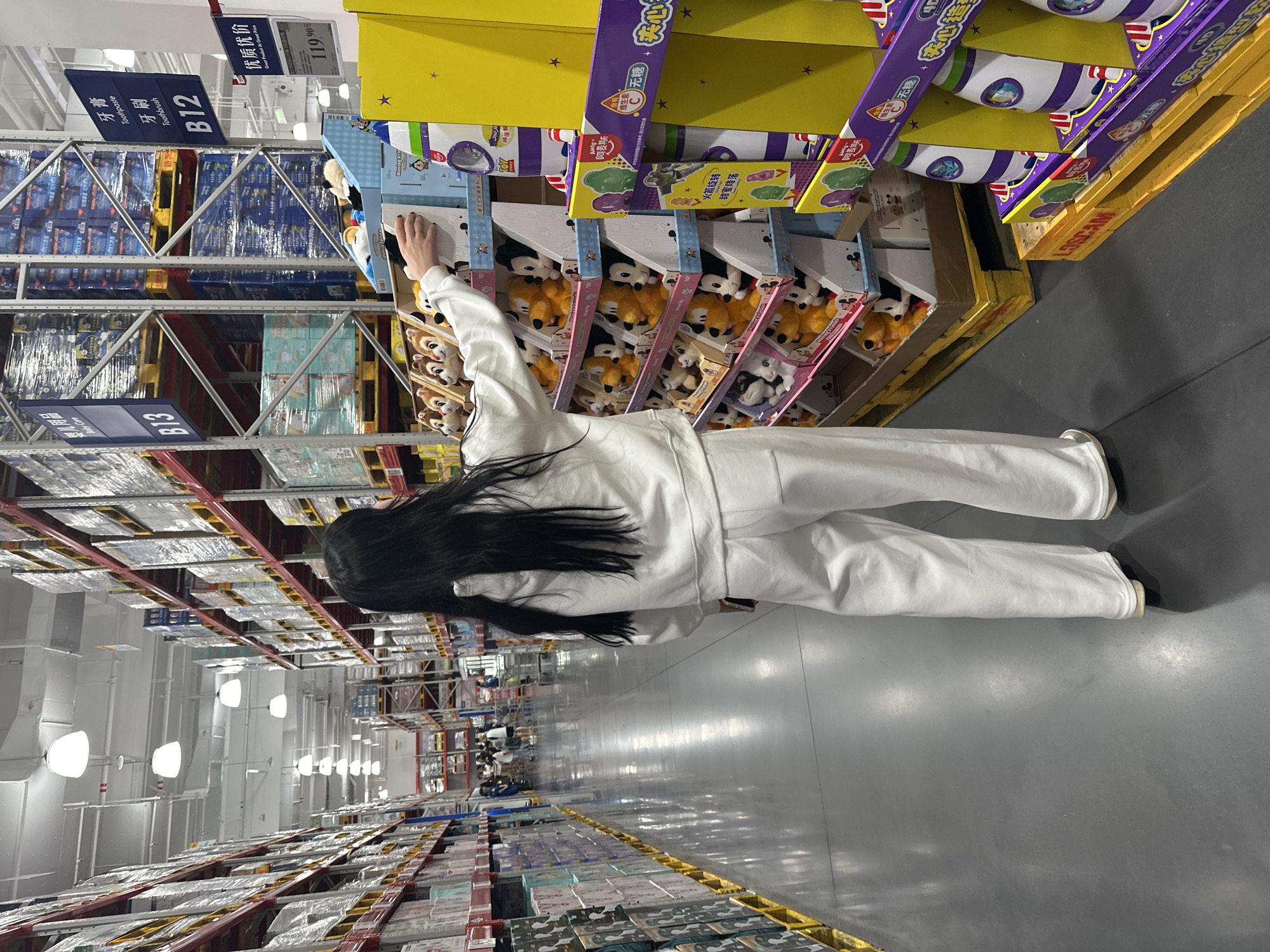 A young woman with long dark hair, wearing a white hooded sweatshirt and white sweatpants, stands in a warehouse-style store, reaching towards a display of plush toys. The store features tall shelving units stacked with boxes of merchandise, including what appear to be diapers and toothpaste. The woman is facing away from the camera, and her right hand is extended towards the toy display. The floor is a polished concrete surface, reflecting the overhead lighting. In the background, other shoppers are visible further down the aisle. The signage on the shelves includes Chinese characters and product names such as "Toothpaste" and "Toothbrush." The overall impression is a retail environment, likely a large discount store or a wholesale club. The image is taken from a slightly elevated perspective, looking down the length of an aisle.