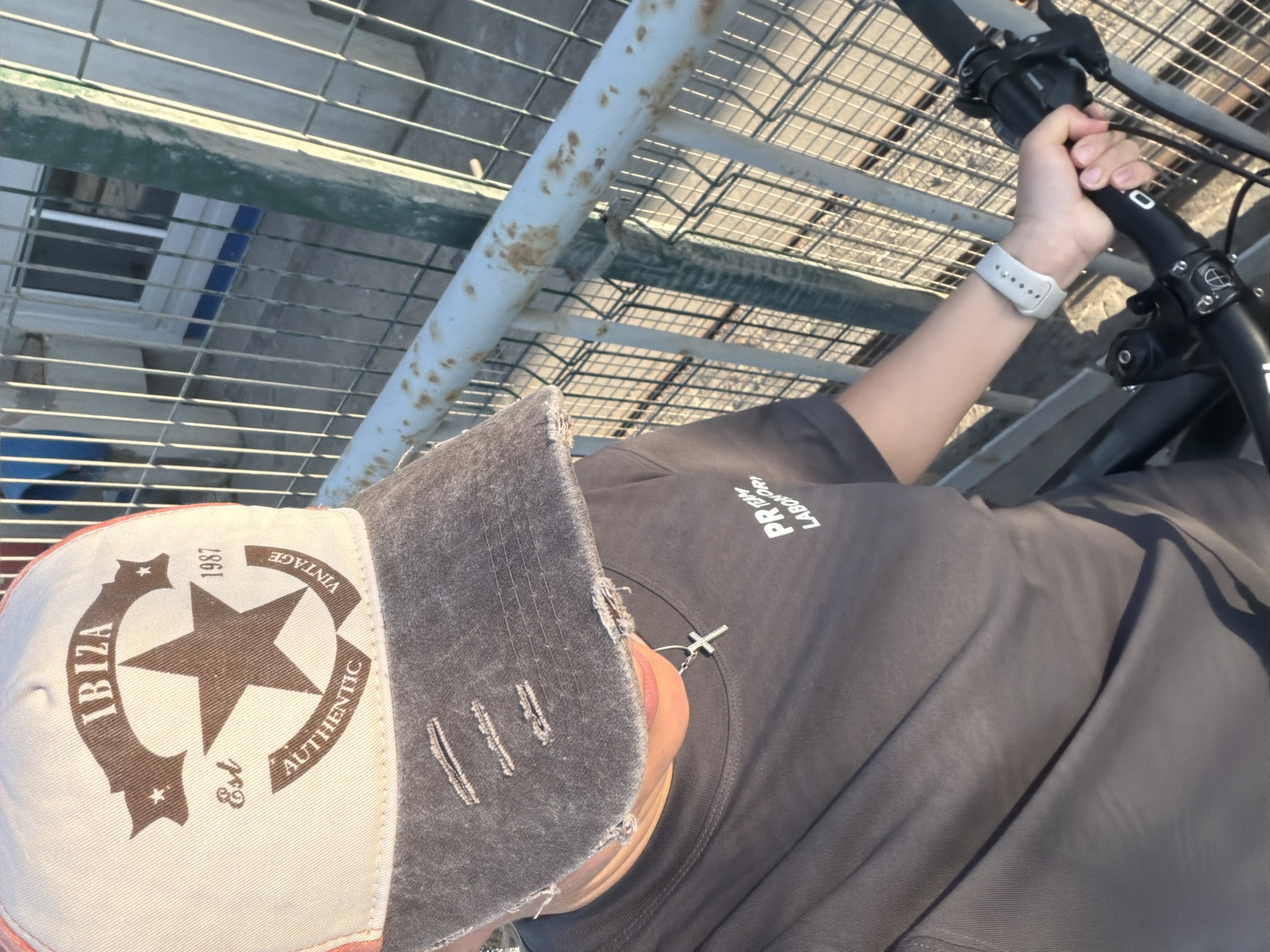 A person is seen from a high angle, holding the handlebars of a bicycle. They are wearing a gray t-shirt with white text on the left side and a baseball cap with "IBIZA" and a star logo. A white smartwatch is visible on their left wrist. They are standing on an elevated metal grate platform, likely a bridge or walkway, overlooking an urban environment with buildings and infrastructure visible below. The location is Chaikovsky, Russia.