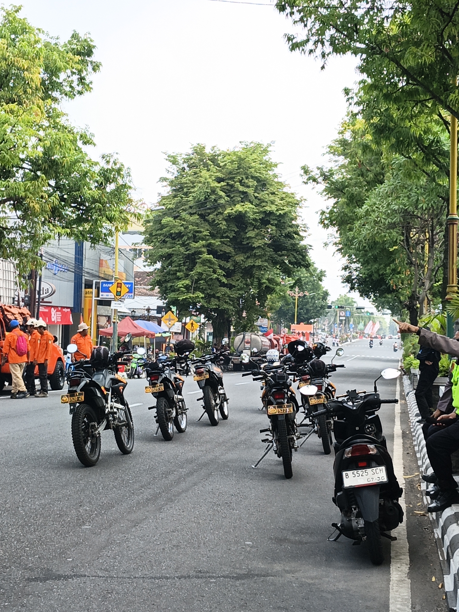 The image depicts a street scene in Bareng, Indonesia, with a line of parked motorcycles on the right side of the road. Several individuals, some wearing orange uniforms, are visible on the left side of the road, near what appears to be a truck. The street is paved, and there are trees lining both sides of the road. Buildings and signs are visible in the background, indicating an urban environment. One person in the foreground is pointing towards the distance. The sky is overcast.