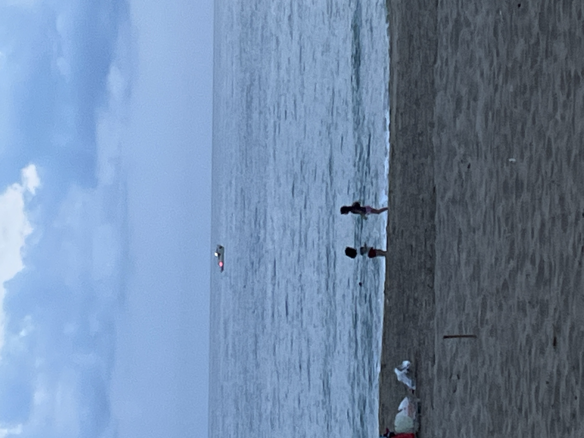 The image depicts a coastal scene, likely a beach, in Alanya, Turkey. The view is vertically oriented, with the sky and clouds occupying the left portion of the image. The sea stretches across the center, leading to a sandy beach on the right.
Two individuals are standing in the shallow water near the shoreline. Their activities or interactions are not clearly discernible due to the distance.
On the beach, several objects are scattered, including what appears to be a white object and possibly a person lying on the sand.
In the distance, a small red and white object is visible on the water. Its identity and purpose are not readily apparent from the image.