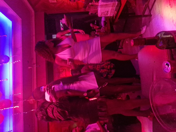 Two women are dressed in light-colored dresses, one white and one appearing pink. They stand close together in a room with vibrant pink and blue lighting. Decorations are visible, possibly balloons, suggesting a celebratory event. A wine bottle is prominently featured in the foreground, enhancing the festive atmosphere. The room appears crowded, implying a social gathering.
