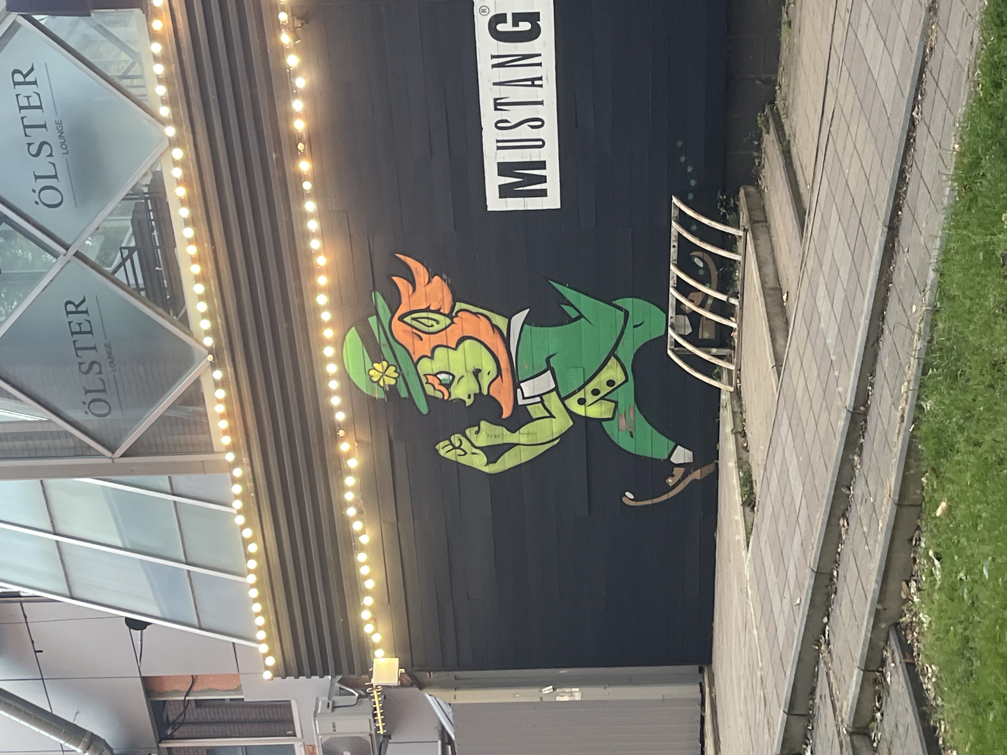 The image shows a graffiti of a leprechaun on the black wall of a building. The leprechaun is depicted in green attire, with orange hair and a green hat adorned with a four-leaf clover. To the right of the graffiti, a white rectangular sign with black lettering reads "MUSTANG". Above the wall, strings of yellow lights are visible, and further up, the building has a modern facade with glass panels and a sign that reads "ÖLSTER LOUNGE". In the foreground, a paved walkway with concrete steps leads away from the building, and a bicycle rack is visible. This scene is located in Krasnodar, Russia.