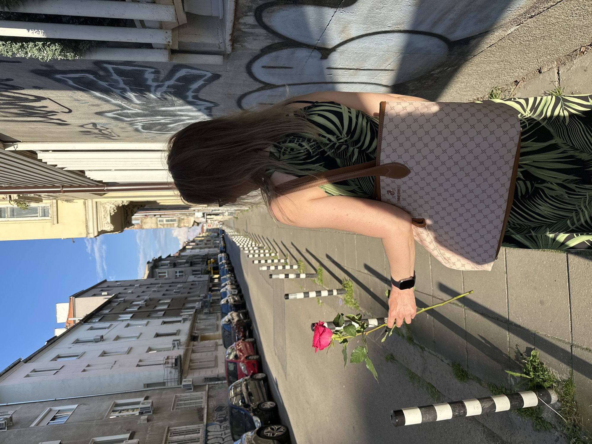 A woman stands on a sidewalk in Sofia, Bulgaria, holding a single red rose. She is viewed from behind, her body turned slightly to the right. She wears a black and green tropical-print dress and carries a light-colored tote bag with a repeating brown logo pattern. A black smartwatch is visible on her left wrist. She is holding the rose in her right hand, close to her body.
The sidewalk is paved with concrete slabs and bordered by black and white striped bollards. Green grass and weeds sprout between the paving stones and around the bollards. The street runs parallel to the sidewalk and is lined with parked cars. Buildings with multiple stories and balconies line the street, some with air conditioning units visible. The walls of one building are covered in graffiti. Sunlight casts long shadows across the scene, indicating it is late afternoon. The sky is blue with scattered clouds.