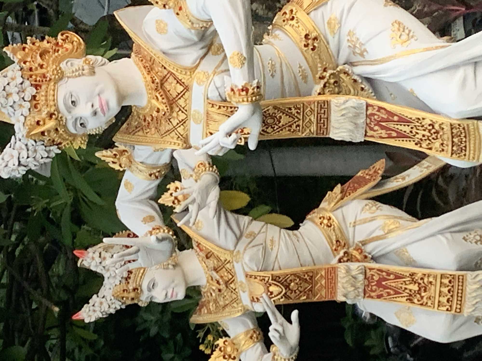 The image displays two white, gold-adorned statues of dancers in traditional Balinese attire. The statues are positioned outdoors amidst lush green foliage. Both figures wear elaborate headdresses, ornate gold jewelry, and flowing white garments with gold trim and red accents. Their hands are posed in a traditional mudra, a symbolic hand gesture. The statue on the left is partially visible, with its head, torso, and arms in frame, while the statue on the right is more fully depicted, showing its entire upper body and lower torso. The background consists of out-of-focus green leaves and branches, suggesting a garden or natural setting.