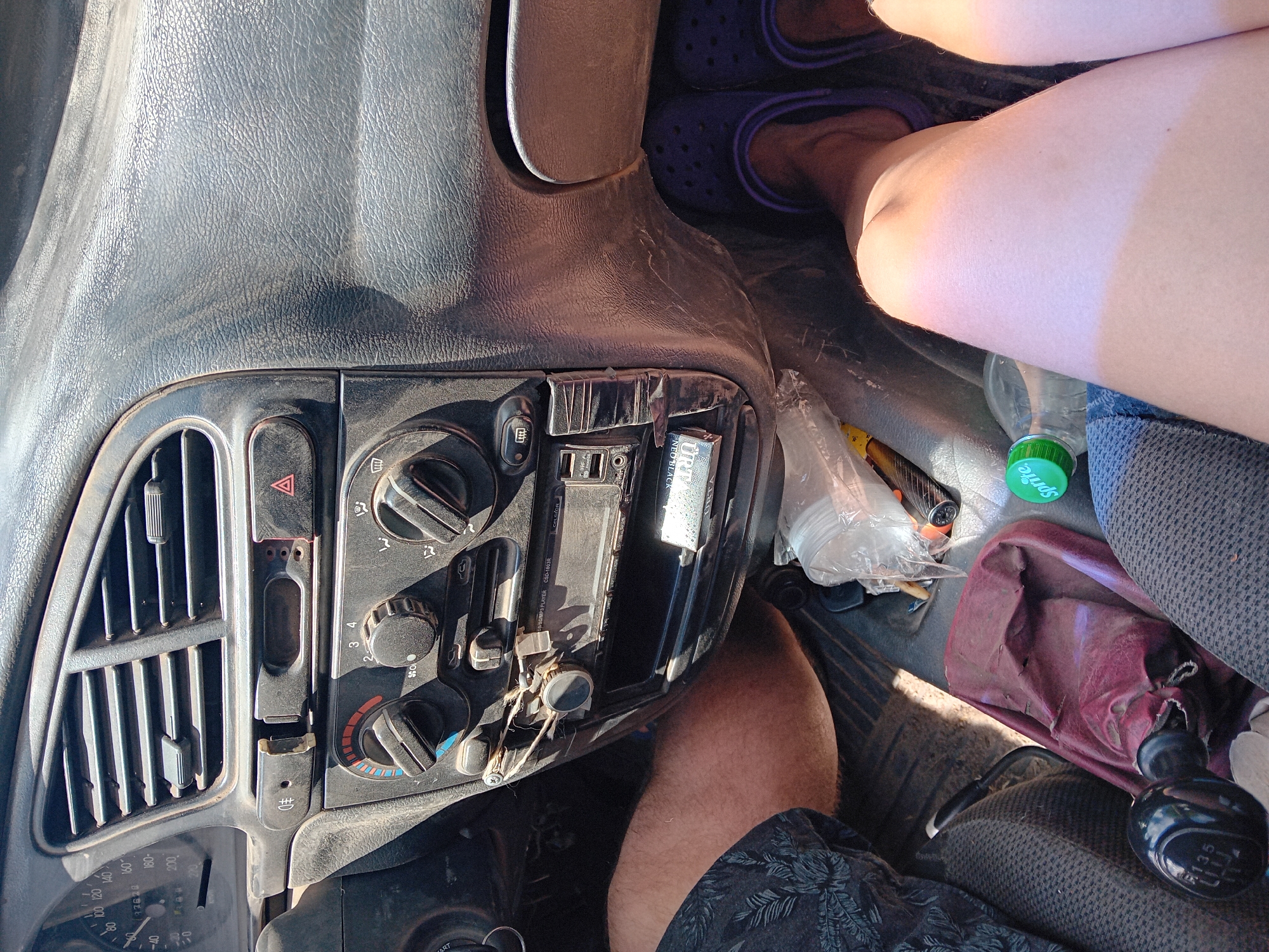 The image shows the interior of a car, likely a Daewoo Lanos, based on the dashboard design, parked in Chuhuiv, Ukraine. The dashboard features a speedometer, air vents, and controls for the climate system. Two individuals' legs are visible, one wearing patterned shorts and the other wearing purple Crocs. A Sprite bottle and a torn maroon fabric are on the floor. The car's gear shift is also visible. The overall scene suggests a casual, everyday situation within the car.