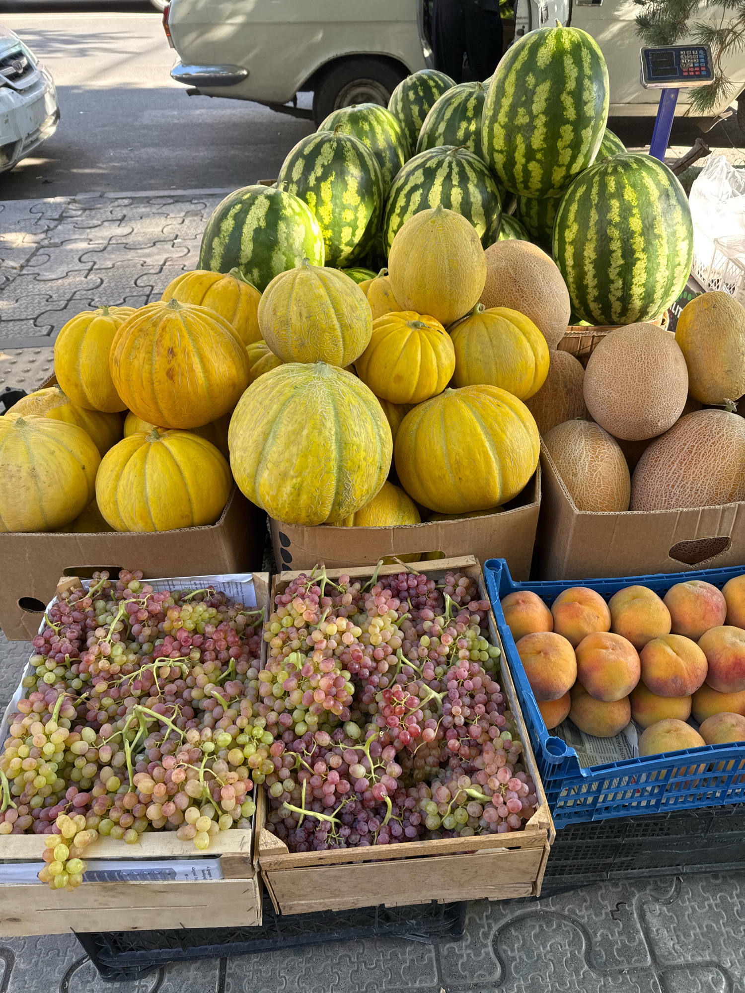 A street vendor's stall displays a variety of fresh fruits, including watermelons, melons, peaches, and grapes. The stall is set up on a sidewalk, likely in a city, with a road and parked cars visible in the background. The fruits are neatly arranged in cardboard boxes and plastic crates, suggesting they are for sale. The overall scene depicts a typical outdoor market setting.