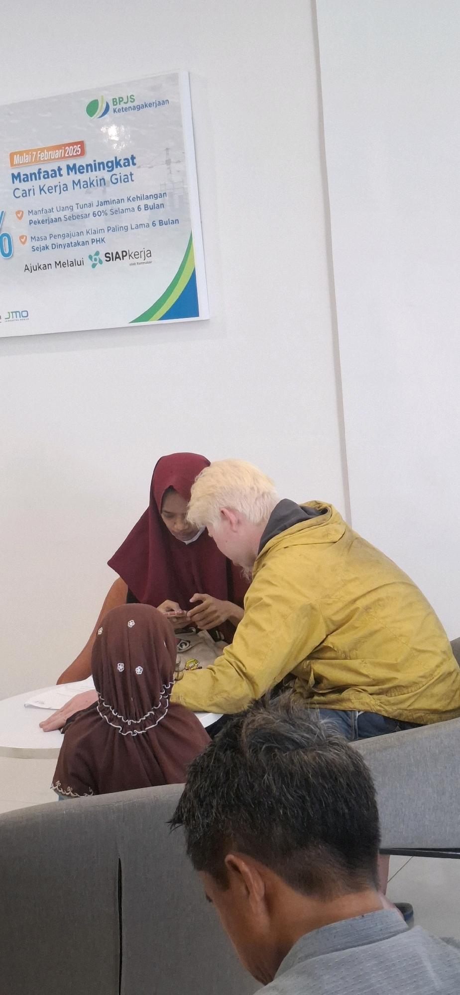 In Surakarta, Indonesia, a group of people are gathered around a table. Two individuals, a woman wearing a maroon hijab and a man with blonde hair, are leaning over the table, seemingly engaged in a discussion or activity. The woman is holding a mobile phone, and the man is looking at it. To their left, a young child wearing a brown hijab with white floral patterns is also present, facing away from the camera. The table is white, and a document or pamphlet is visible on it, featuring the text "Manfaat Meningkat Cari Kerja Makin Giati" and the logo of "BPJS Ketenagakerjaan." In the foreground, the back of another person's head, with short, dark, textured hair, is visible, suggesting they are seated. The overall environment appears to be an indoor waiting area or office, with a neutral-colored wall in the background and a grey sofa on the right. The lighting is bright, indicating an overhead light source.