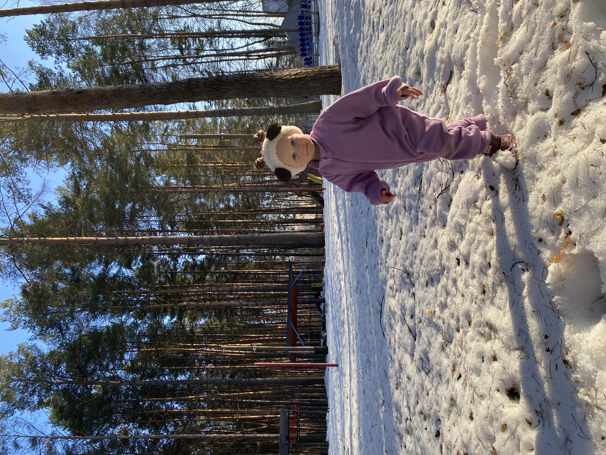 A young child wearing a purple jumpsuit and a panda-themed hat stands in the snow. The child is positioned slightly off-center in the image, looking at the camera. The background shows tall pine trees on the left side, creating a forest-like environment. In the distance on the left, there is a partially visible playground structure. The ground is covered with snow, and fallen pine needles are scattered on the snow surface. The image was taken in the city of Chaikovsky, Russia.