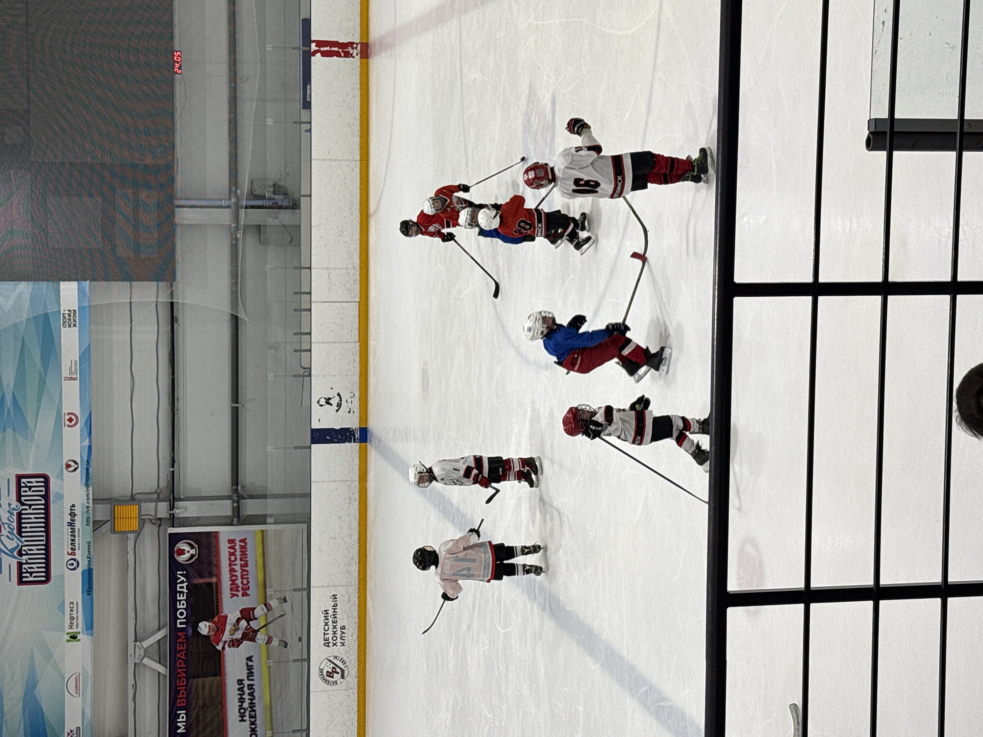 The image shows a hockey game taking place at an indoor rink. Several young hockey players wearing protective gear and holding hockey sticks are on the ice. They wear various team colors including red, white, and blue. A digital clock displays "24.05." Banners with Russian writing and logos, including the logos of the hockey club and sponsors, are visible along the walls. The location is likely an arena in Votkinsk, Russia, based on the Russian text and sponsor affiliations on the banners.