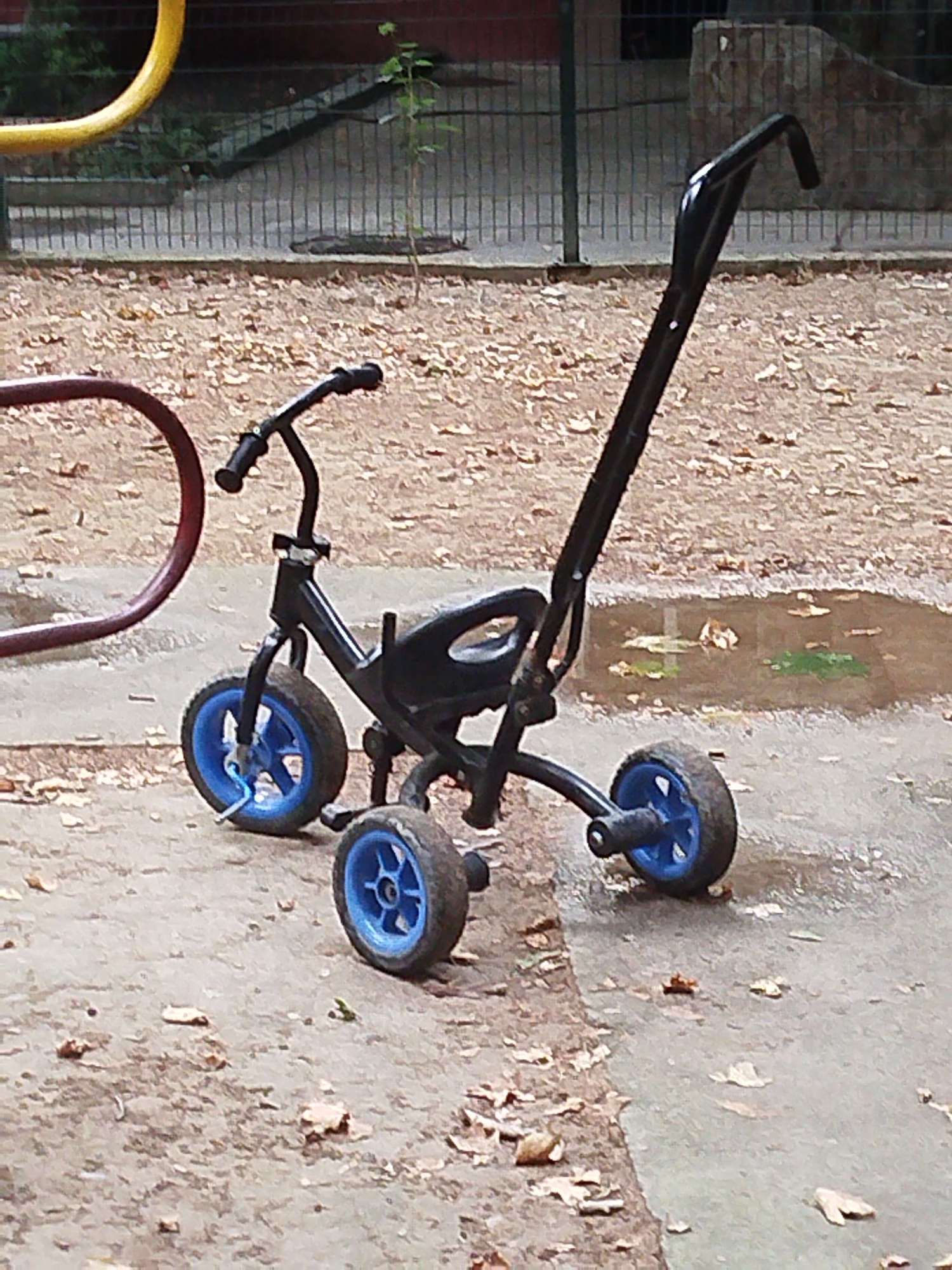 A black tricycle with blue wheels is parked on a wet, sandy surface dotted with fallen leaves. The tricycle has a push handle, a seat, and three wheels. One of the rear wheels appears to be in a puddle. In the background, a blurry yellow bar from playground equipment is visible, along with a metal fence and some greenery. The scene suggests an outdoor setting, possibly a park or playground, after a rain. No people are visible, and there is no discernible text in the image.