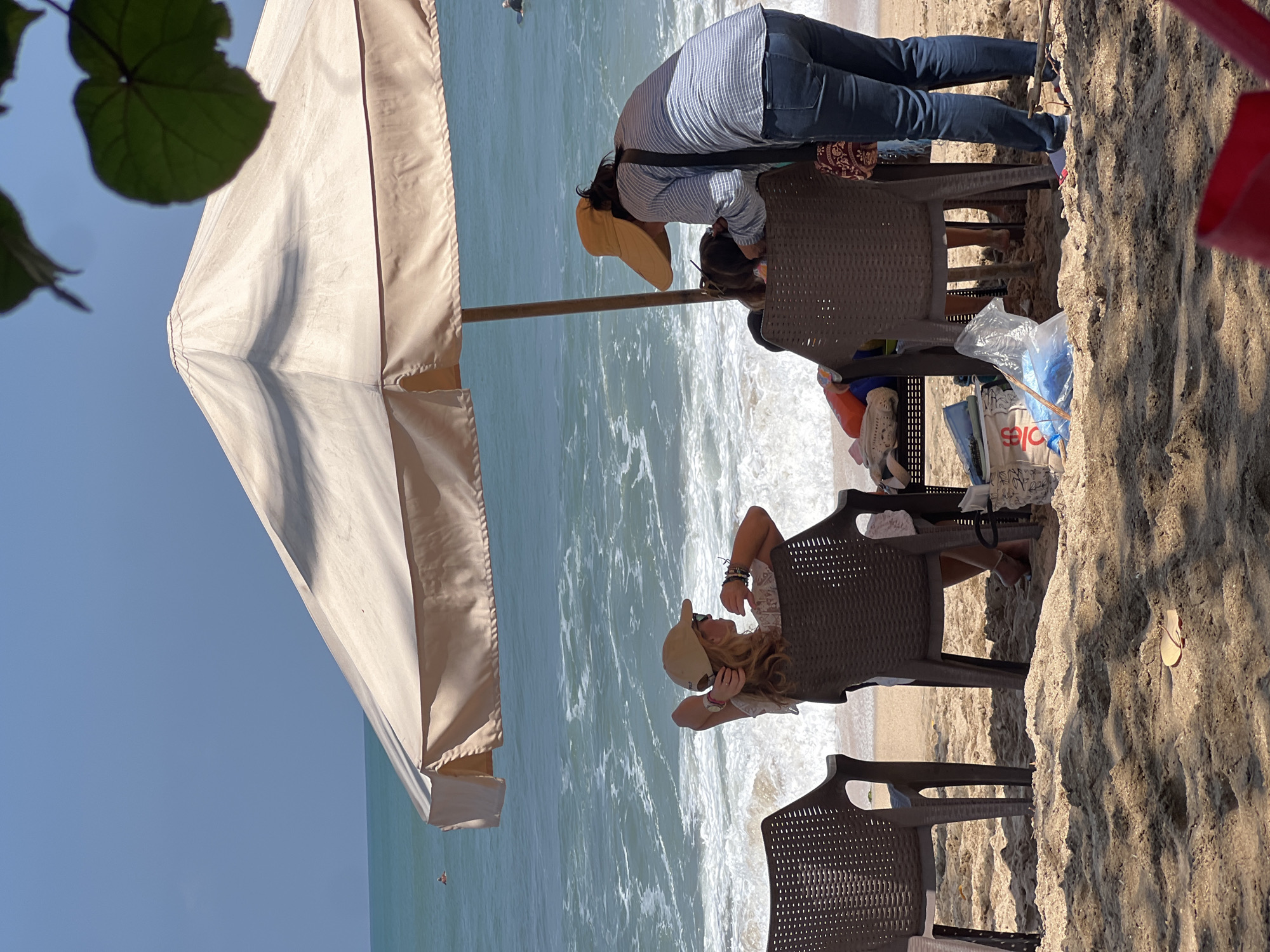 Two women are seated under a large, cream-colored beach umbrella on a sandy beach. One woman, wearing a beige baseball cap, is seated in a brown plastic chair facing the ocean, with her hands behind her head. The second woman, wearing a wide-brimmed yellow hat, is standing and leaning over, interacting with someone seated to her right. Several brown plastic chairs and a table are present under the umbrella. In the background, the ocean has moderate waves, and the sky is clear and blue. The sandy beach shows footprints and scattered debris, including a red flip-flop. This scene is consistent with a typical beach setting in Kuta, Indonesia, known for its popular tourist beaches and umbrella rentals.