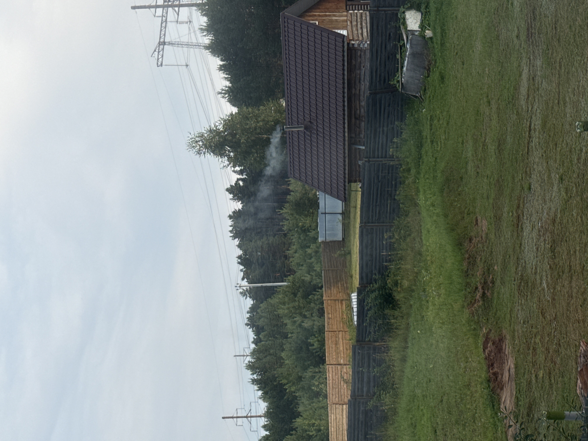 A wooden cabin with a dark brown, shingled roof is situated in a rural setting, likely on the outskirts of Chaikovsky, Russia. Smoke billows from the chimney of the cabin, indicating activity within. A dark wooden fence runs horizontally across the foreground, separating the grassy yard from the area behind it. Beyond the fence, a dense forest of pine trees stretches into the distance. In the background, high-voltage power lines and pylons are visible against a cloudy sky. The overall scene depicts a tranquil, rustic environment.