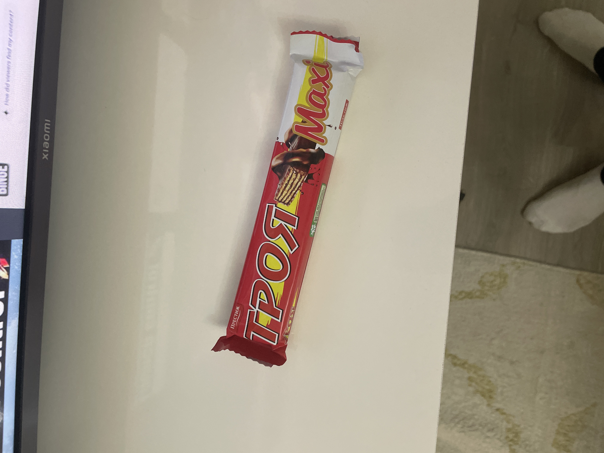 An indoor, eye-level photograph captures a packaged chocolate bar resting on a light-colored, possibly white, surface. The chocolate bar is primarily red with yellow and white accents, featuring the brand names "Maxi" and "ТРОЯ" (Troya) prominently. An image of a chocolate-covered wafer is visible on the wrapper. Additional Cyrillic text, "ПРЕСТИЖ" (Prestizh), is also visible on the packaging.
To the left of the frame, a portion of a digital display screen is visible, oriented vertically. The brand "xiaomi" is clearly identifiable, along with the word "BINGE" and the question "How did viewers find my content?". This suggests the screen is likely a television or monitor displaying a content creation or streaming interface.
The right side of the image reveals a floor area, part of which is covered by a light-colored, patterned rug. Two feet, clad in plain white socks, are visible on this rug, suggesting a person is standing or sitting nearby.
The setting appears to be a casual indoor environment, possibly a living room or office in Ihtiman, Bulgaria, given the Cyrillic text on the packaging and the provided location context. The lighting is ambient, without strong shadows to indicate a specific time of day. No specific activity is occurring other than the presence of the objects and the person's feet.