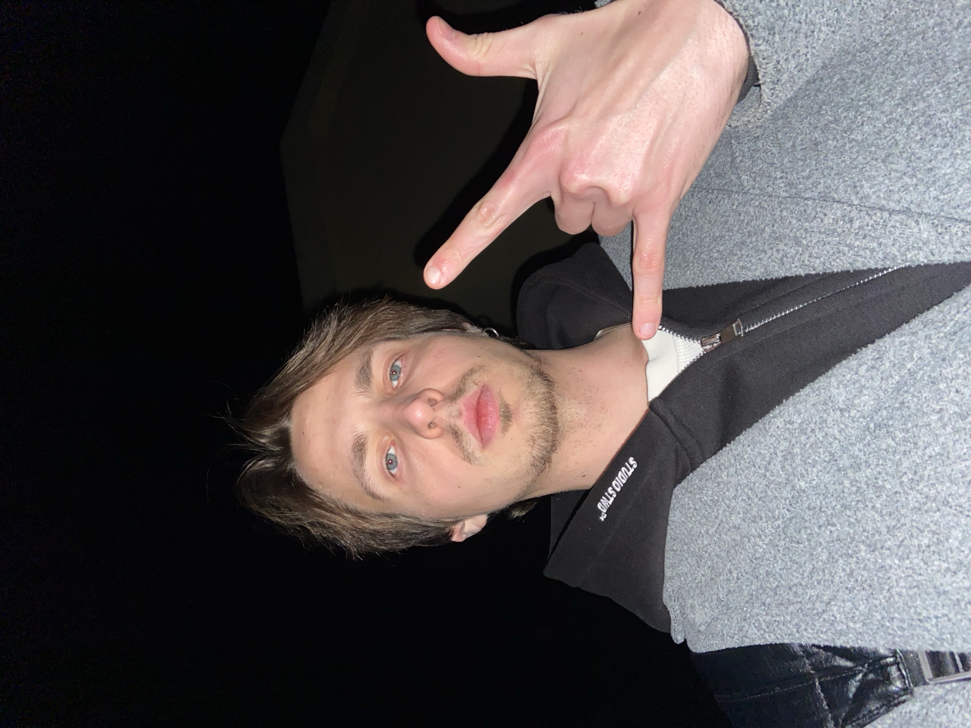 A man with light brown hair, a trimmed beard, and blue eyes is positioned in the center, looking up at the camera. He is wearing a black zippered sweatshirt with white lettering on the collar and a grey coat. His right hand is raised above his head, forming an L shape. The background is dark, with a faint indication of a structure in the upper left. Based on the location prompt, this photo was taken in Šušanj, Montenegro.