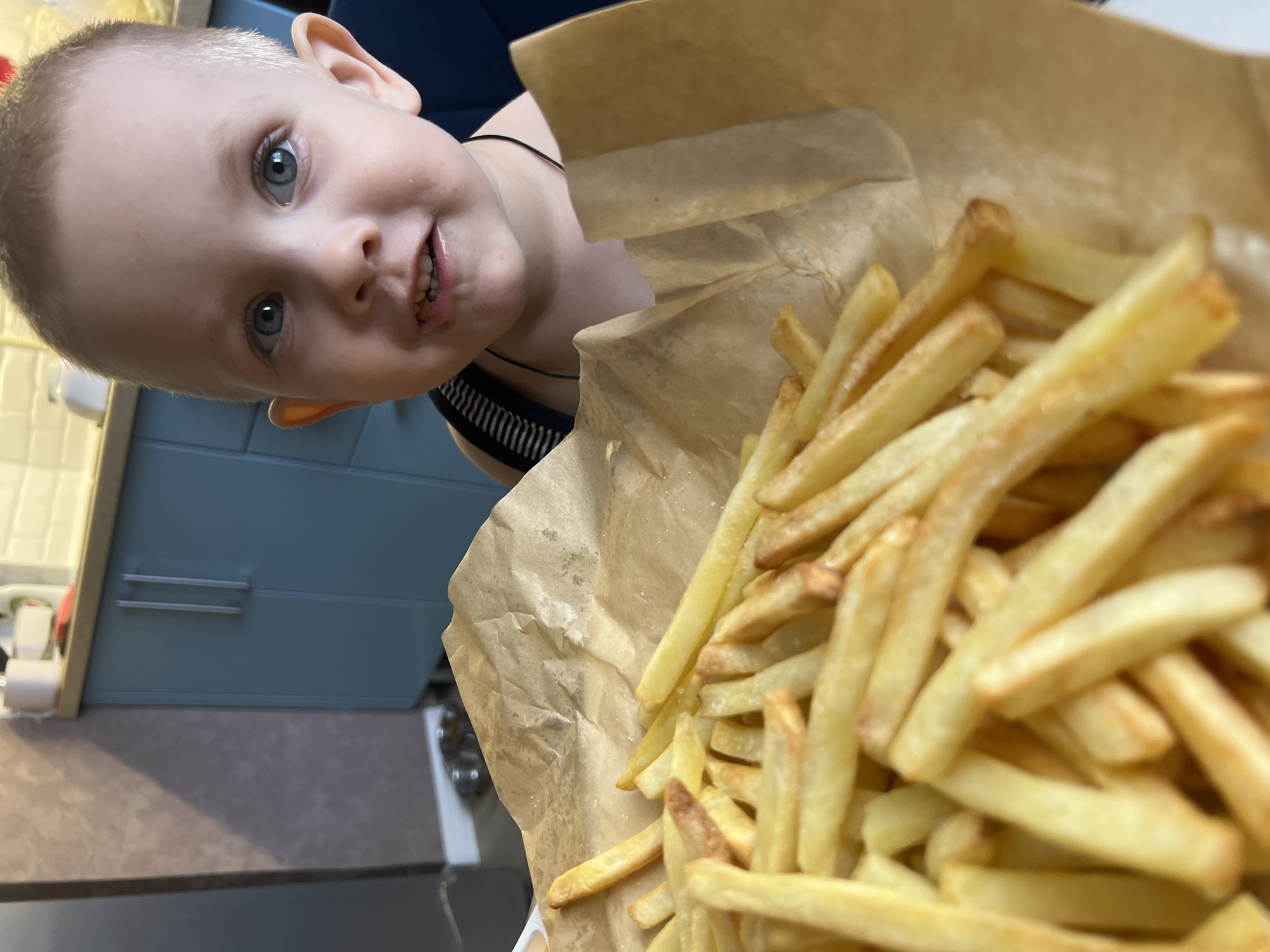 A young child with short blonde hair and blue eyes, wearing a black and white striped tank top and a black choker necklace, is looking directly at the camera with a slight smile. The child is positioned to the left of a pile of french fries served in a brown paper bag. The fries are light golden yellow and appear to be seasoned.
In the background, there is a blue cabinet with silver handles, a counter surface, and a white object which appears to be a roll of paper towels. The setting appears to be indoors, likely a kitchen. The specific location is Марковский, Russia.