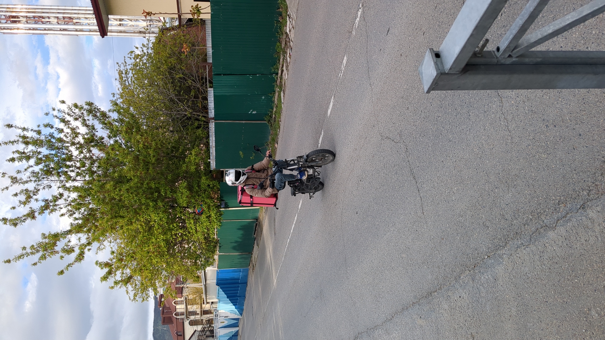 A delivery driver on a small motorcycle with a red delivery box is riding on an asphalt road marked with a dashed white line. To the left of the road is a green metal fence and a tree. In the background are buildings and a utility structure. The scene is in Gelendzhik, Russia. A metal structure is visible in the upper right corner. The sky is blue with clouds.