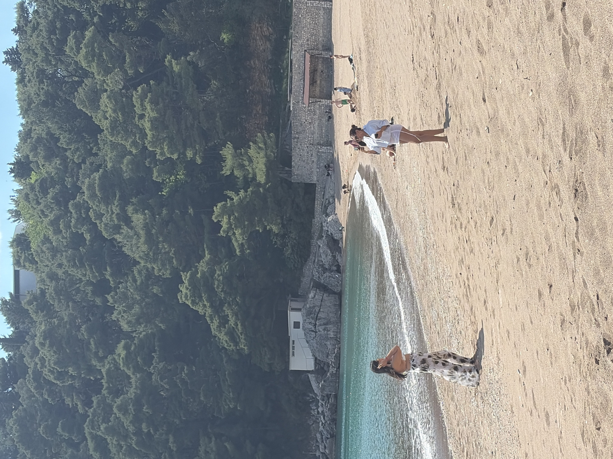 The image depicts a beach scene in Boreti, Montenegro. The beach is sandy and stretches along the coastline. On the left, dense green trees cover a hillside. A group of people is standing on the beach, some near the water's edge. A woman in a light-colored patterned dress stands near the water, and a group of people in white clothing stands further up the beach. The water appears clear. Some buildings and stone structures are located near the beach. Some people are doing yoga exercises in a wooden structure.